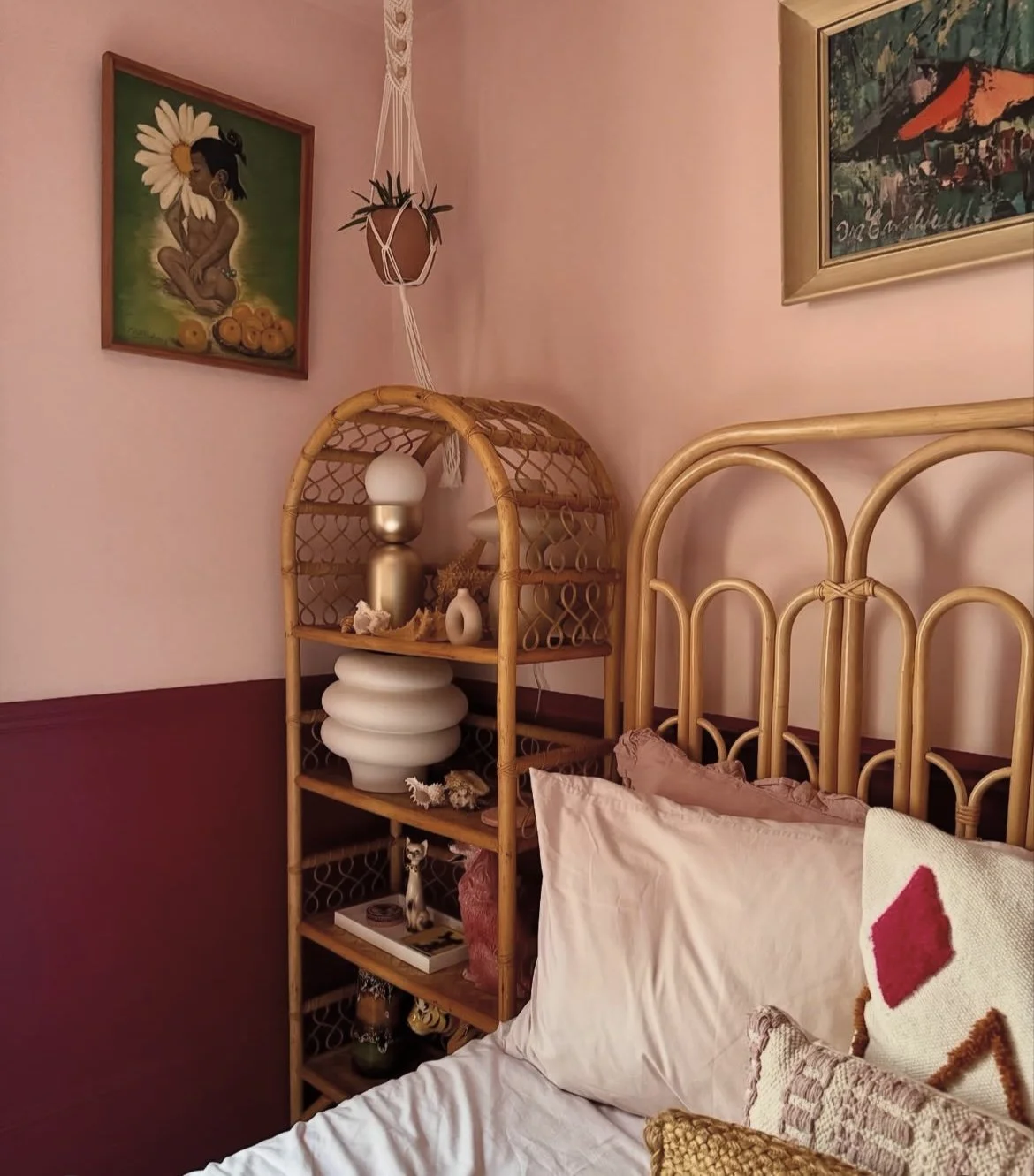 A bedroom corner with a rattan bed frame, a white bed with decorative pillows, a wooden shelving unit with vases and seashells, pink walls, and framed artwork including a painting of a girl with a large daisy and another of tropical scenery.