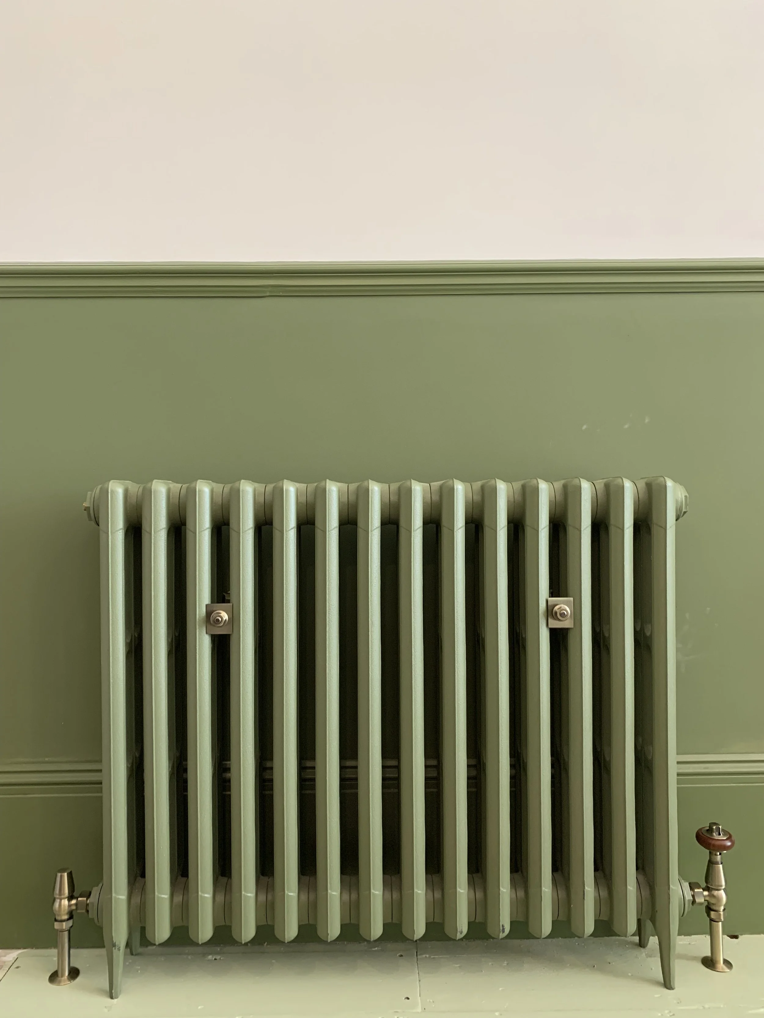 A vintage green cast iron radiator against a green wall with a chair rail, with a floor showing tiles or linoleum.