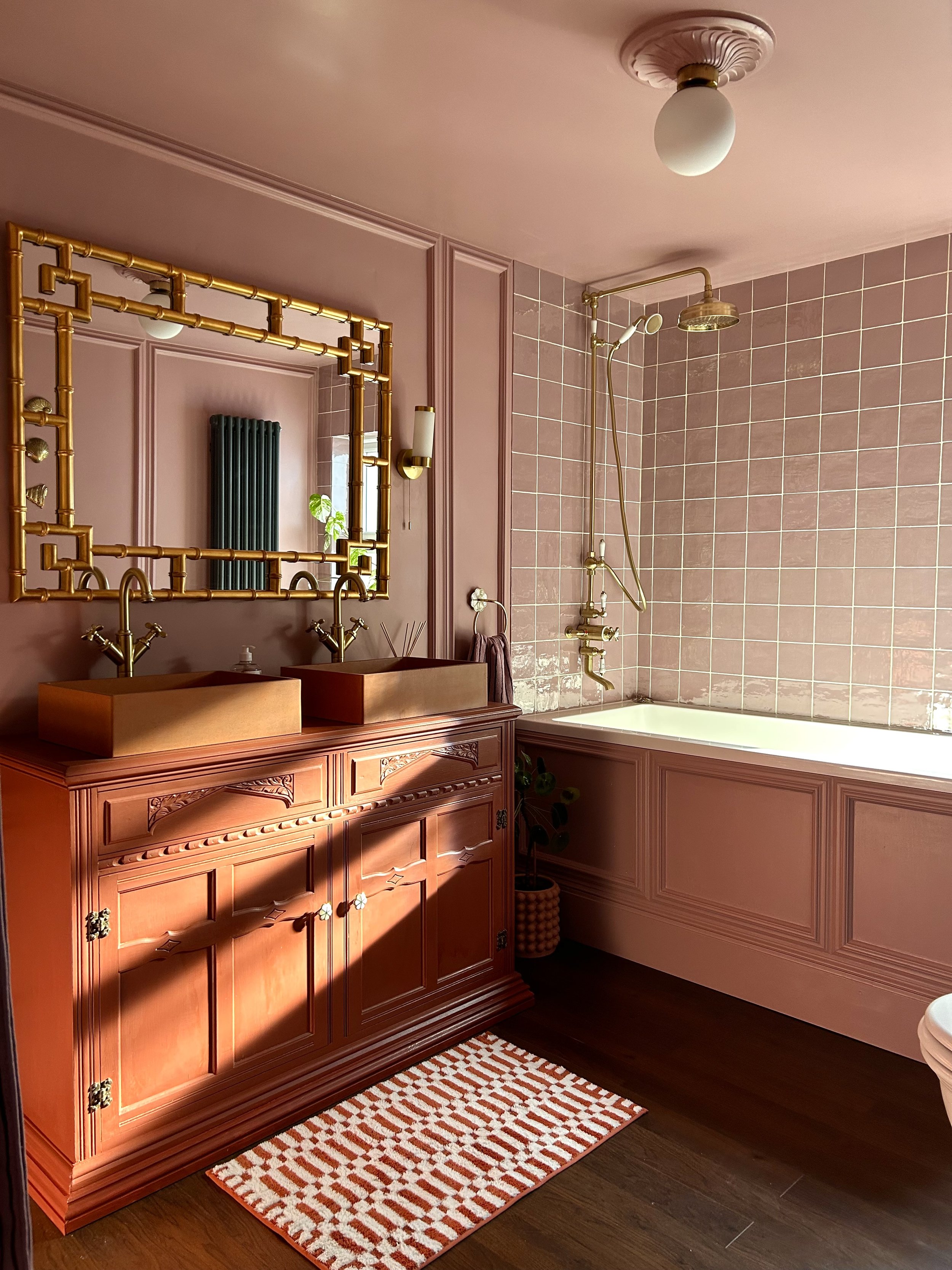 A stylish bathroom with pink walls, a large mirror with a gold bamboo-style frame, two copper sinks, a pink bathtub with a gold showerhead, and a small orange and white patterned rug on a wooden floor.