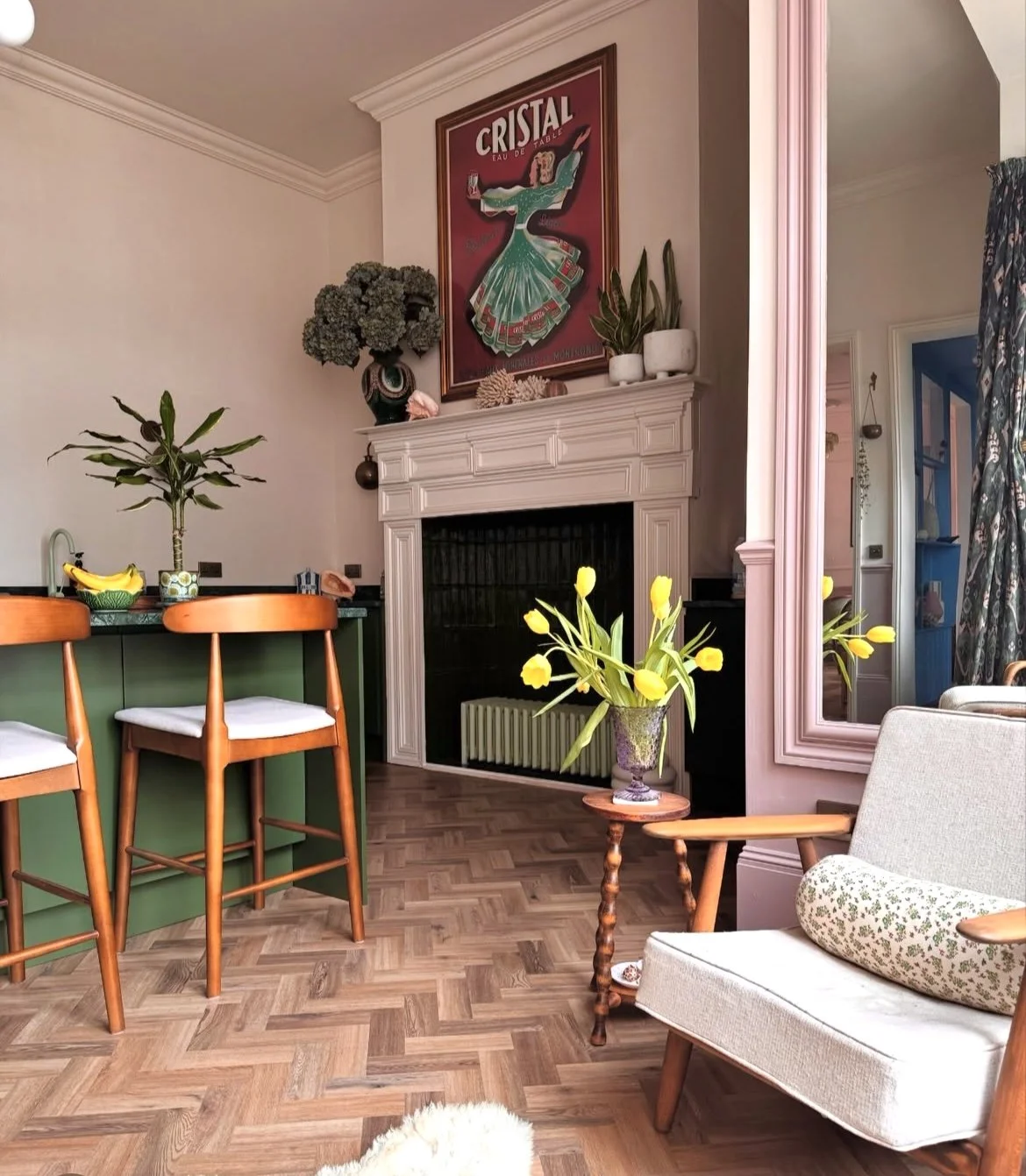 A cozy living room with a white fireplace, a framed vintage advertisement poster above it, a green bar with two wooden chairs, a small table with yellow tulips in a vase, a grey armchair with a floral pillow, and various potted plants, including a banana plant and other greenery, around the room.