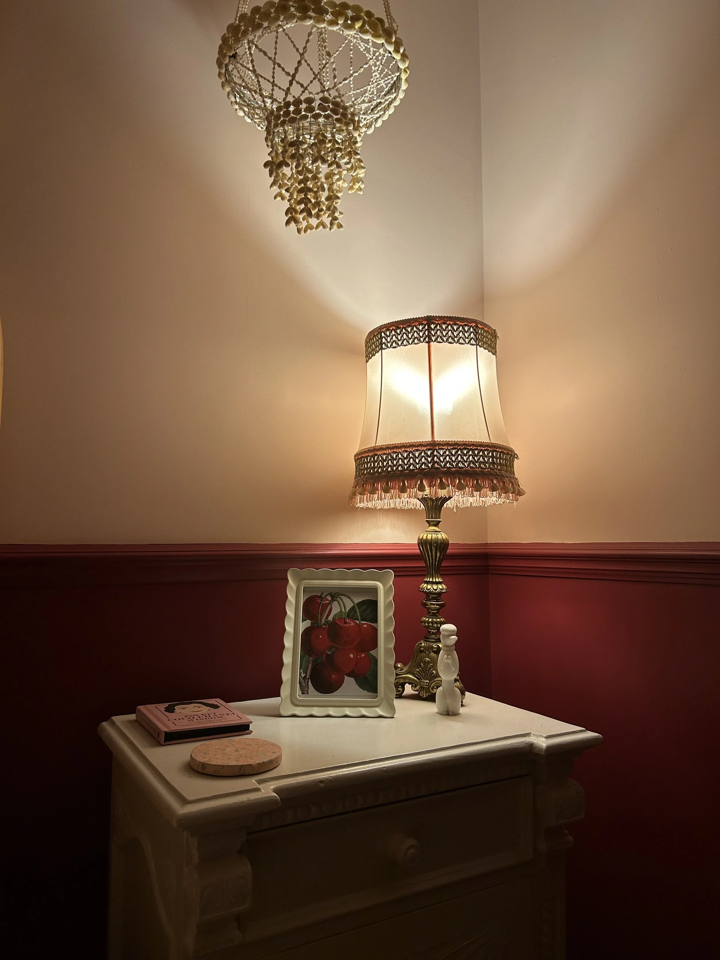 A vintage table lamp with a fabric shade and decorative trim on a white dresser, next to a framed picture of cherries, a small book, and two ceramic figurines, with a beaded chandelier hanging from the ceiling overhead.