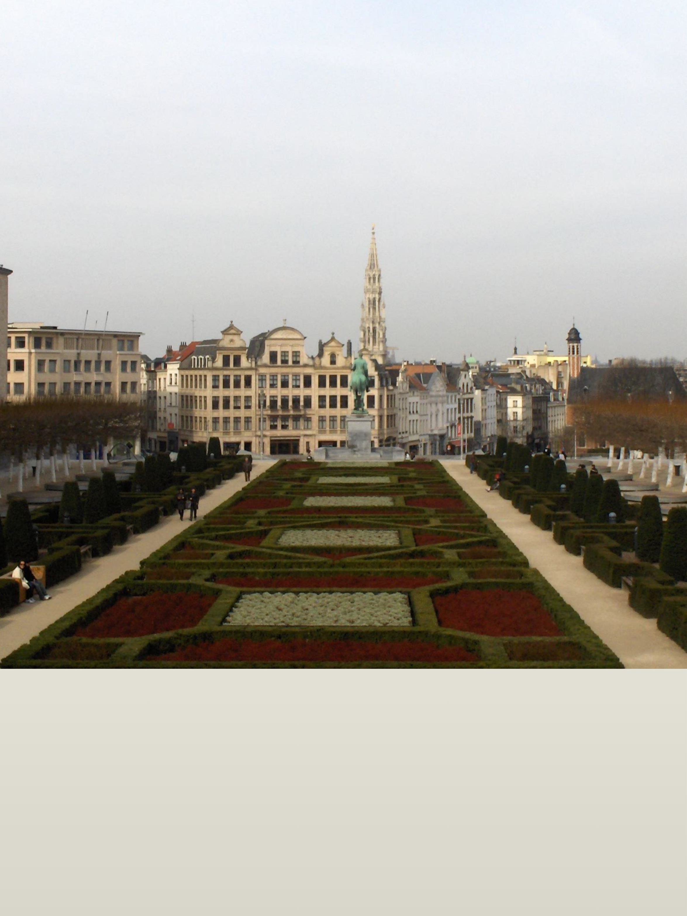 Une vue d'une place avec une statue équestre au centre, entourée de jardins bien entretenus et d'immeubles anciens, avec une église en arrière-plan sur une journée ensoleillée avec un ciel clair.