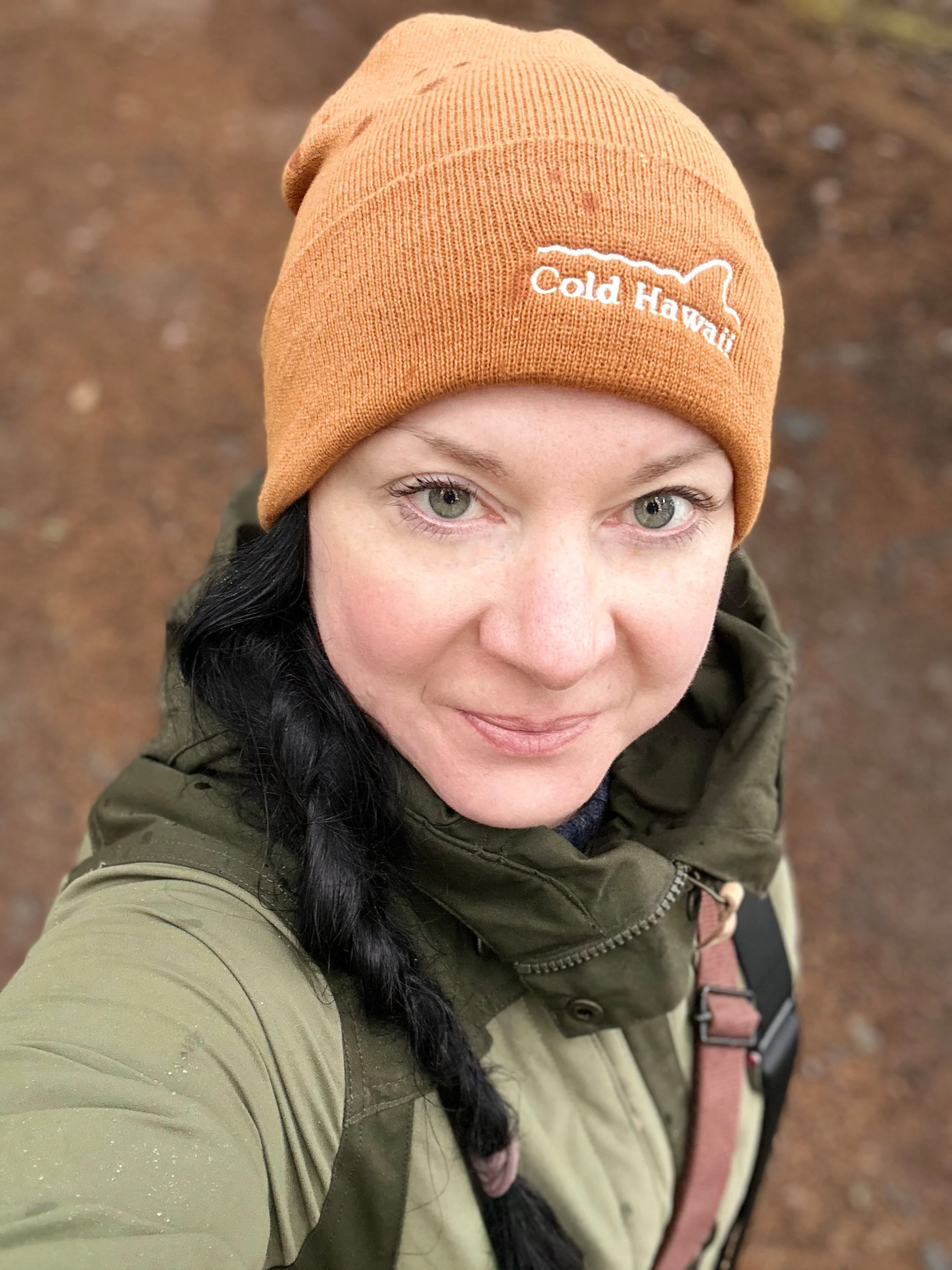 Nahaufnahme eines lachenden Mädchens mit schwarzer Zopf, das einen olivgrünen Mantel trägt und eine braune Mütze mit der Aufschrift 'Cold Hawaii' trägt, im Hintergrund ein erboste, braune Erdfläche.