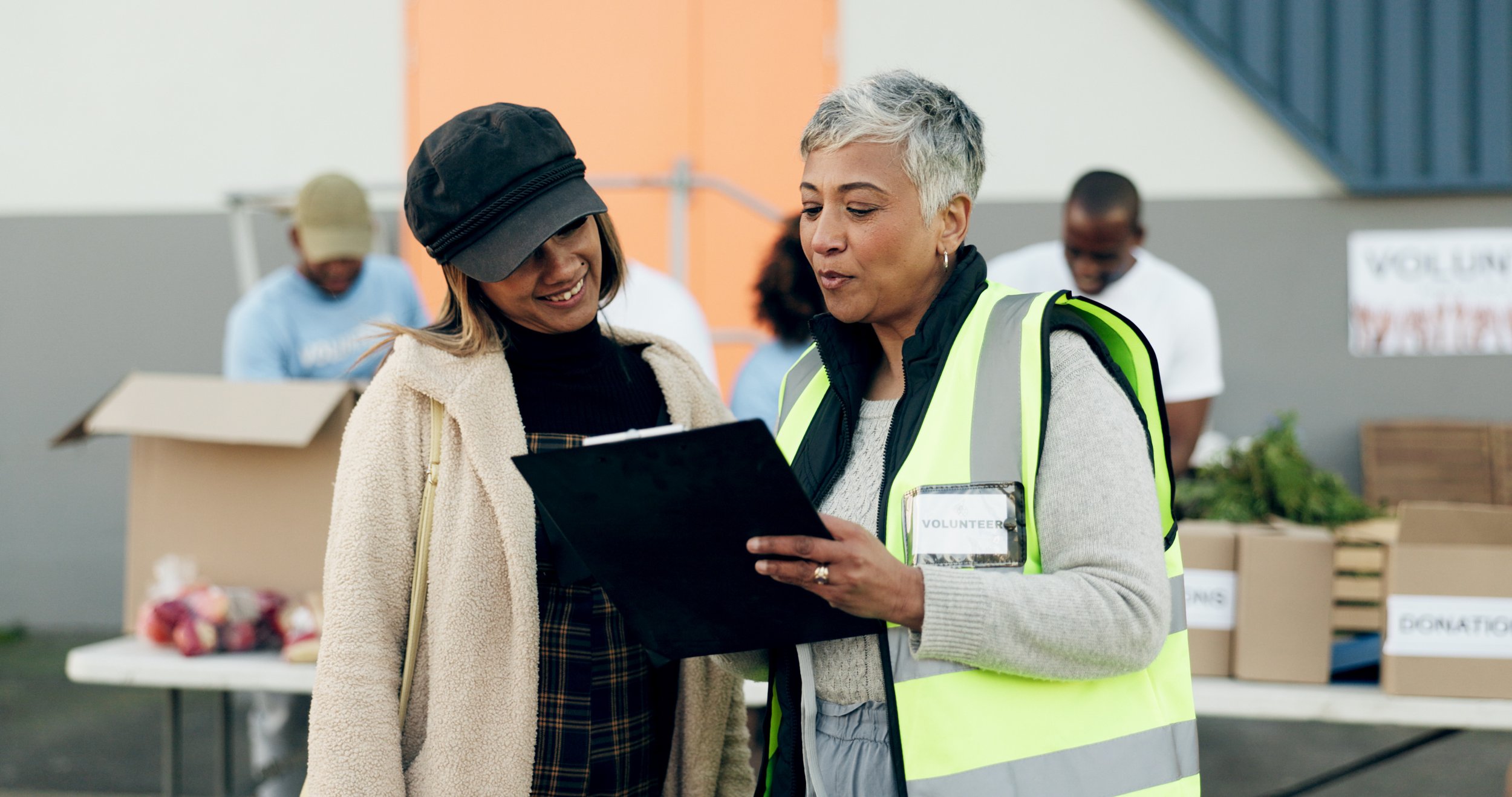 Two women, one wearing a volunteer badge and a reflective vest, looking at a clipboard together, at a food donation event with boxes of supplies in the background.