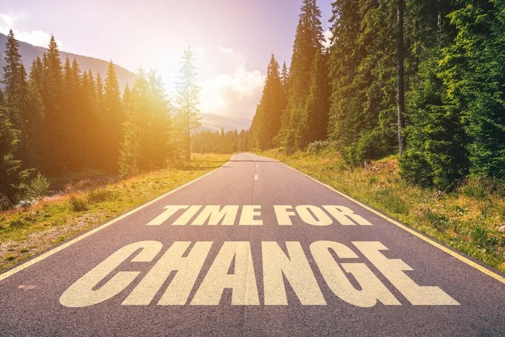 A paved road surrounded by a forest of tall evergreen trees under a partly cloudy sky, with the words 'TIME FOR CHANGE' painted on the road surface.