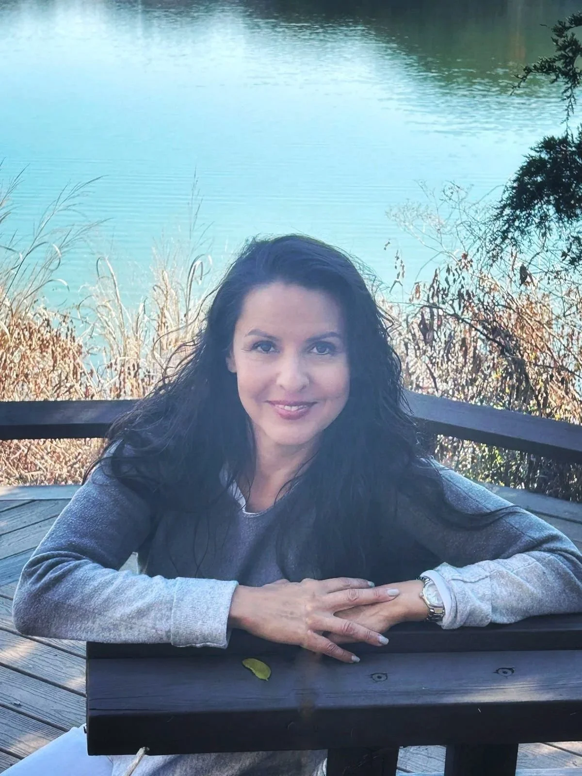 A woman with dark curly hair, wearing a light gray sweater, sitting at a dark wooden table outdoors near a lake with calm water and overhanging trees.