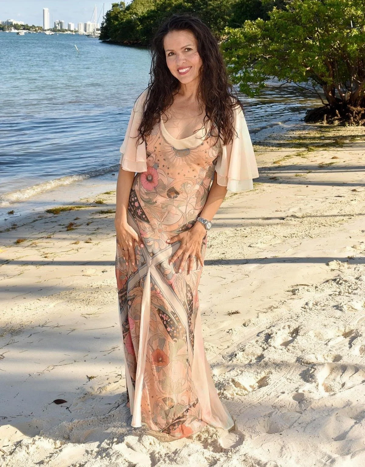 Woman standing on sandy beach on Biscayne Bay, wearing a colorful floral dress with ruffled sleeves, smiling and posing for the camera.