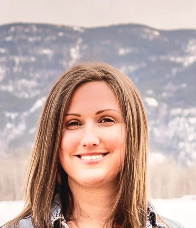 A smiling woman with brown hair standing outdoors with mountains and a cloudy sky in the background.