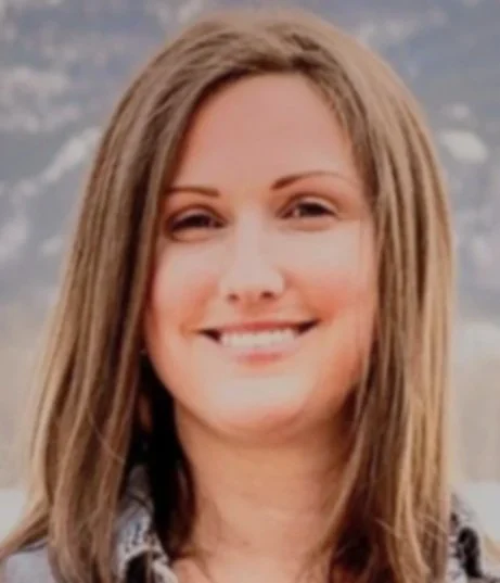 A woman with long brown hair smiling outdoors, with mountains in the background.