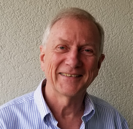 A smiling older man with gray hair wearing a blue and white striped shirt, standing against a beige textured wall.