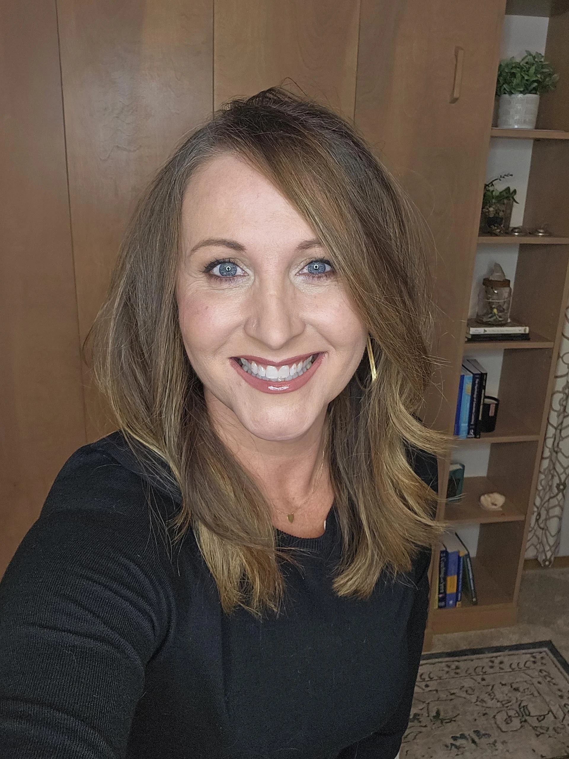 A woman with shoulder-length light brown hair and blue eyes smiling at the camera in an indoor setting with wooden furniture and bookshelves in the background.