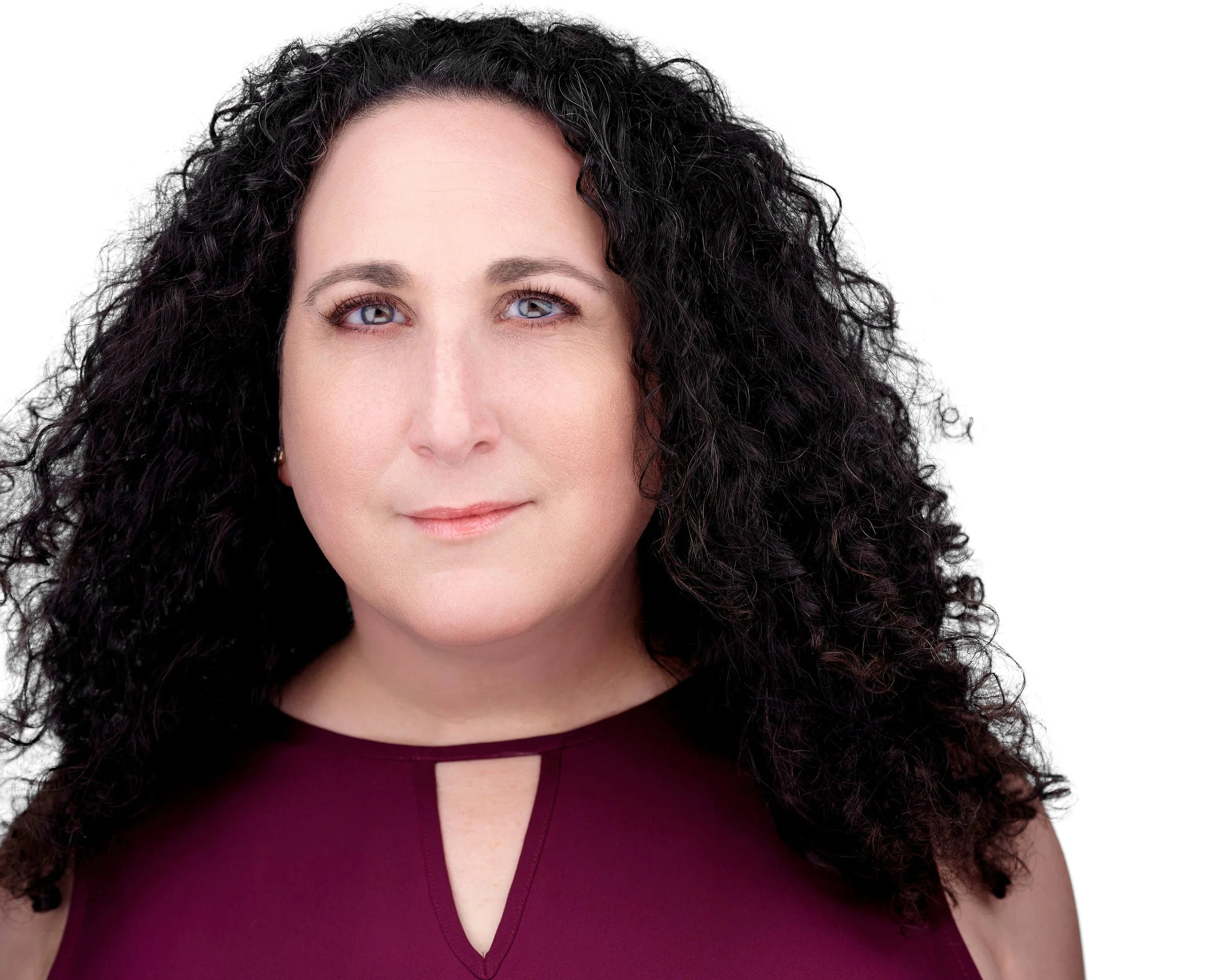 A woman with curly dark hair and light blue eyes wearing a sleeveless burgundy top with a keyhole neckline, posing against a white background.