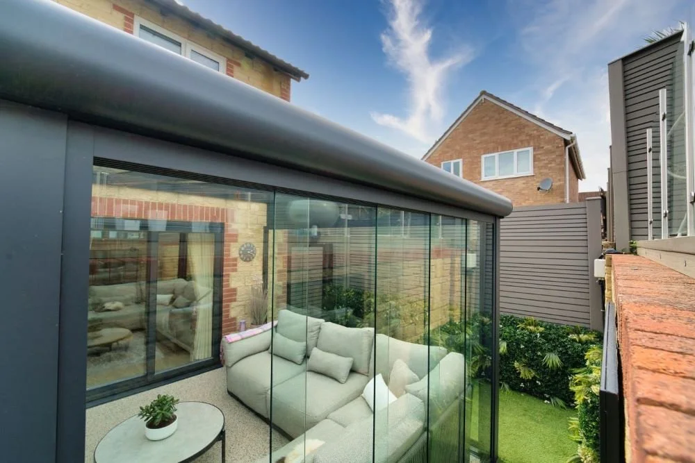 View of a modern glass sunroom attached to a house, with outdoor garden and neighboring houses visible.