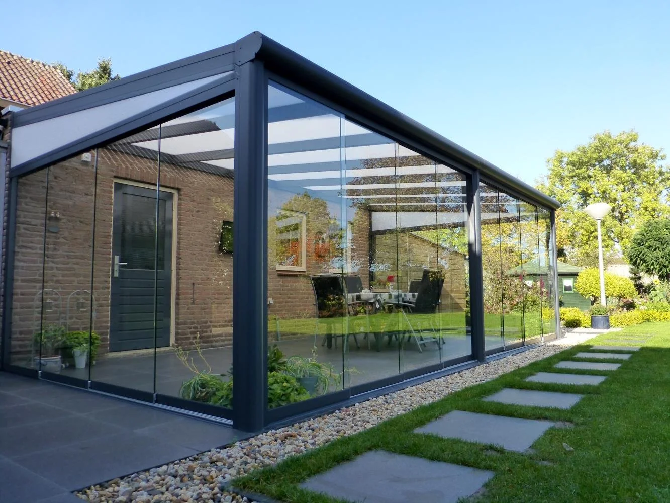 A modern glass extension added to a brick house with a glass roof and paneled walls, surrounded by a well-maintained garden with stepping stones and landscaping.