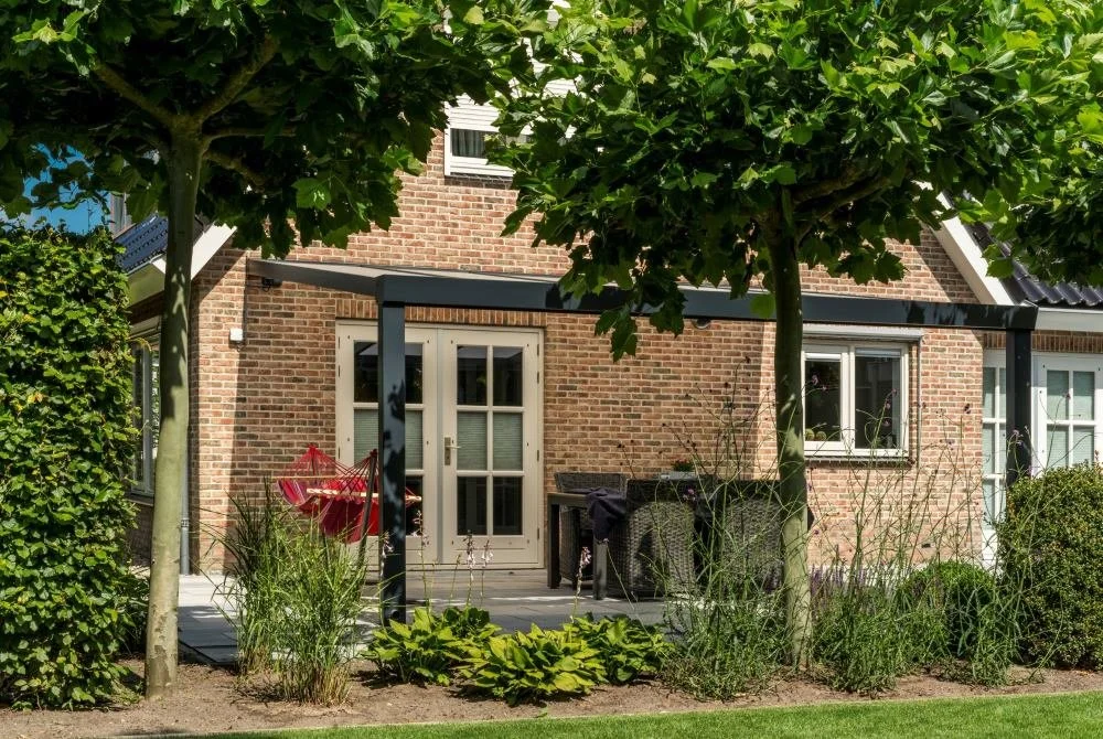 Backyard patio with trees, shrubs, and a garden, attached to a brick house with white doors and windows.