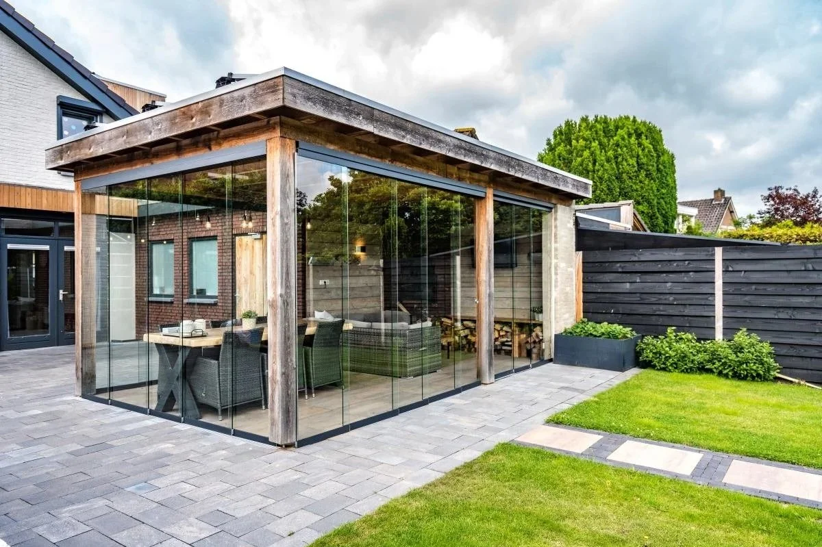 A modern glass-sided patio enclosure attached to a house, with a dining setup inside, overlooking a well-maintained lawn and garden area.