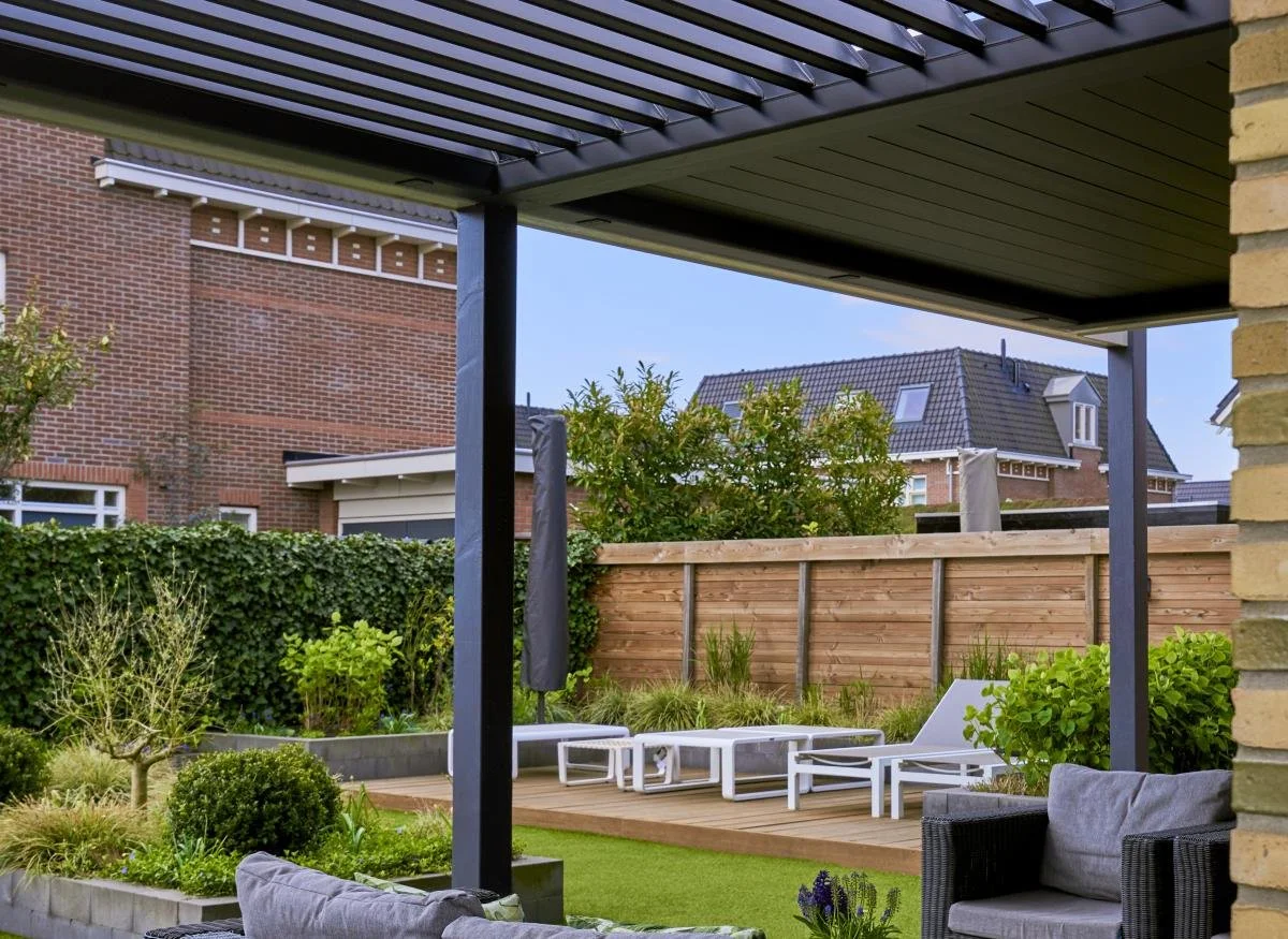 Backyard patio with a wooden deck, lounge chairs, a sofa, and lush greenery, fenced by a wooden fence, with neighboring houses visible in the background.
