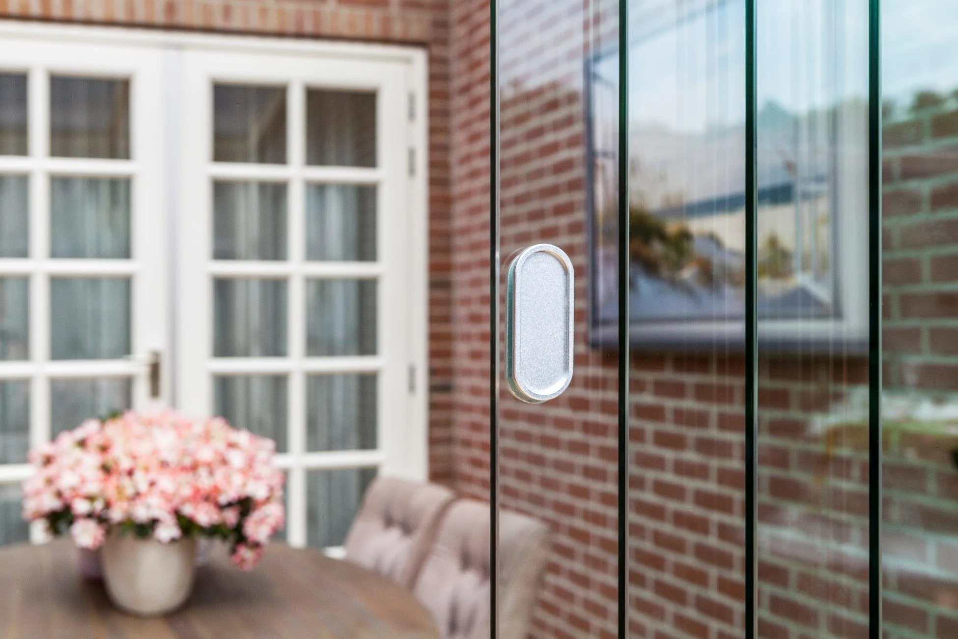 Close-up photo of a modern glass door handle on a dark-framed glass door, with outdoor scene reflected in the glass, and a background showing a brick wall, a white-framed window, a door, a round table with a pink flower arrangement, and a pink cushio