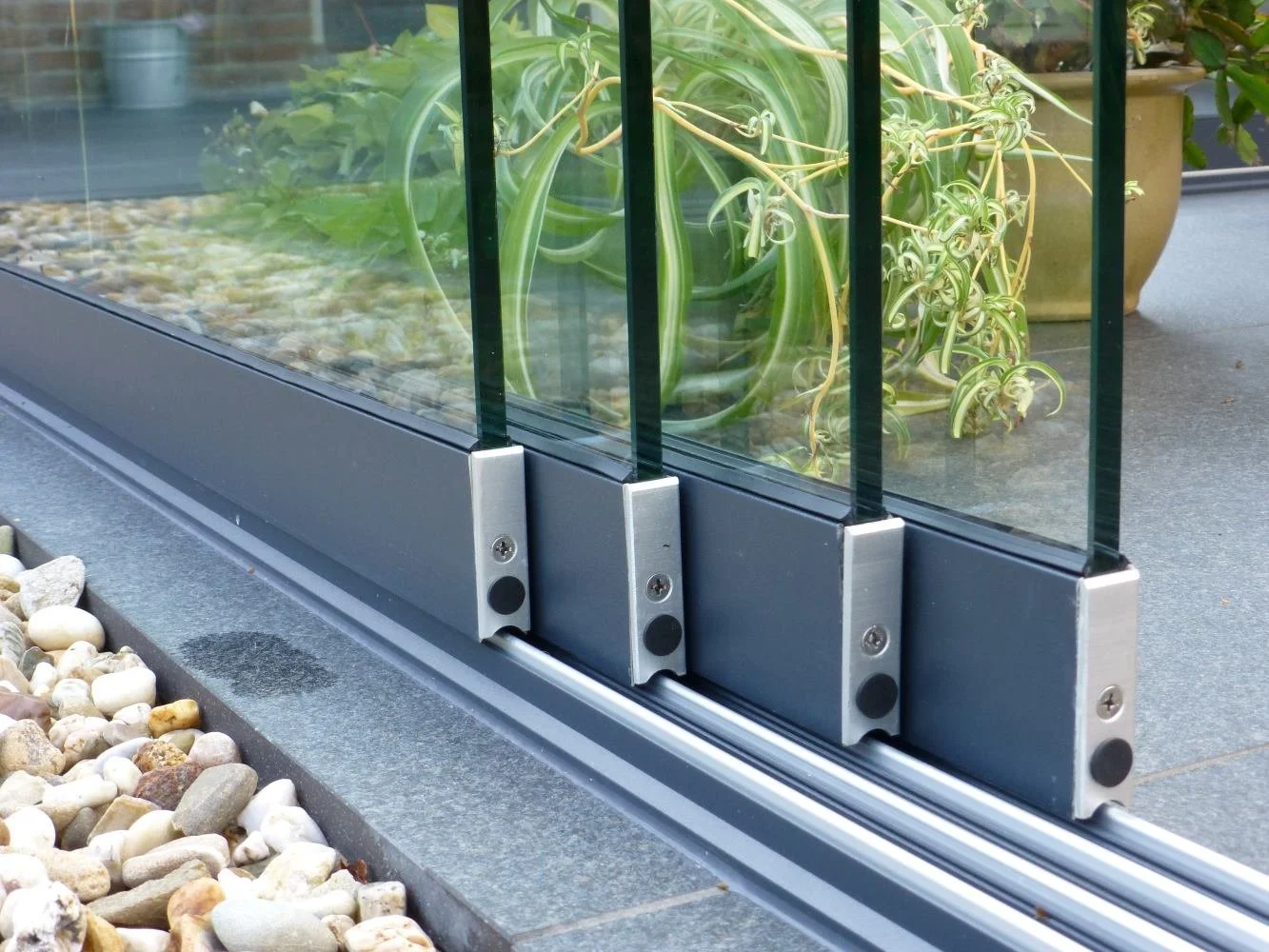 Close-up of glass panels with metal brackets and a sliding track at the bottom, part of a glass fence or barrier, with potted plants and decorative rocks nearby.