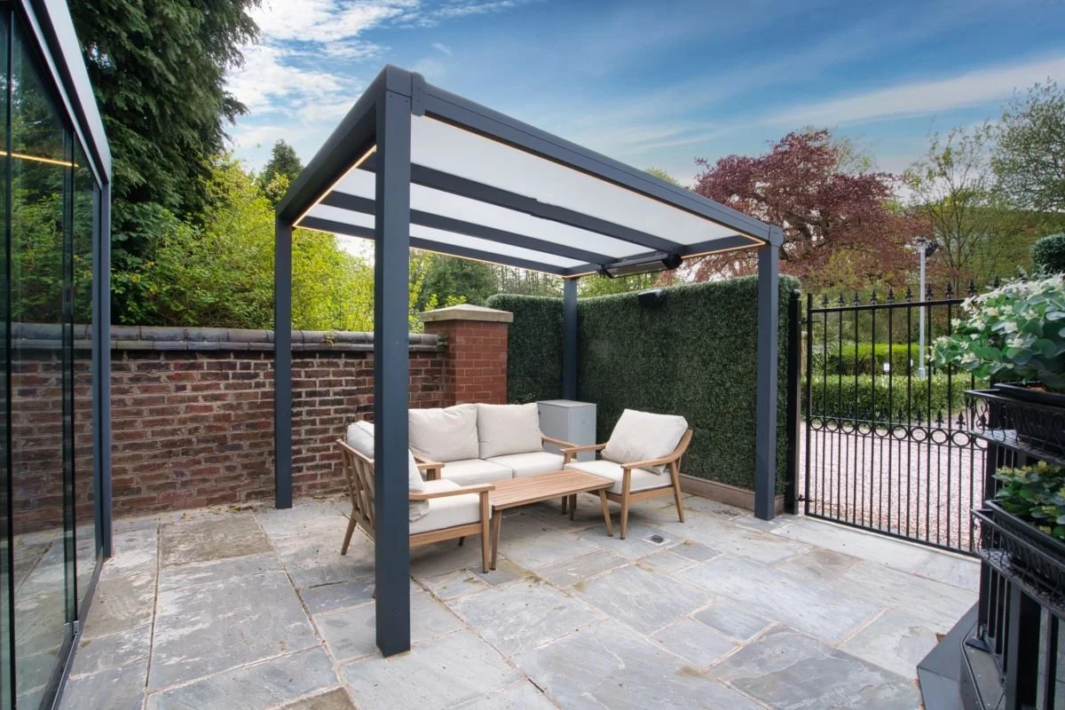 Outdoor patio with a white cushioned sofa and armchair under a modern black pergola, surrounded by greenery, a black metal fence, and brick and glass walls.