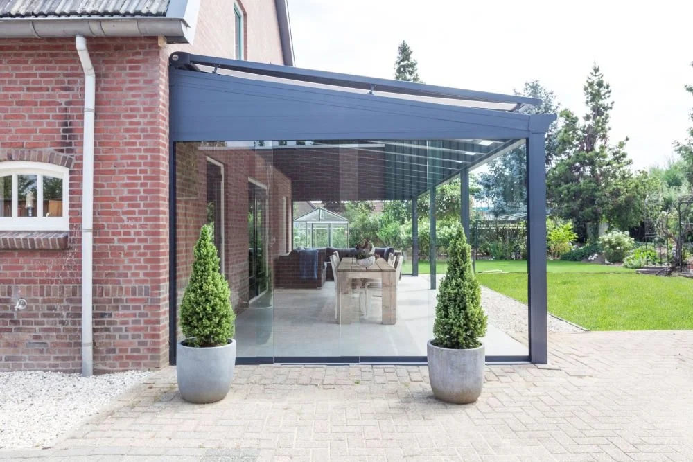 A glass patio enclosure attached to a brick house with potted plants outside, green lawn, and trees in the background.