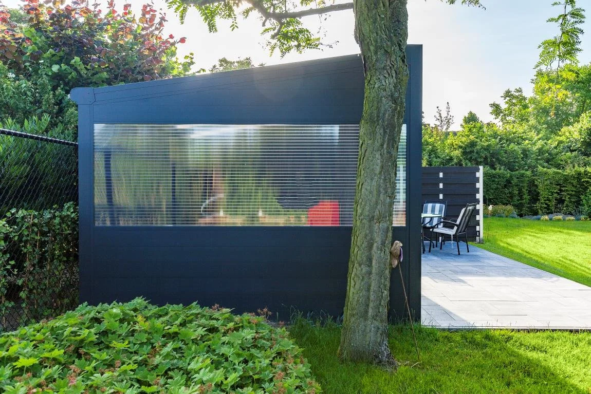 A backyard with a modern dark-colored shed with a translucent window, surrounded by greenery, a tree in front, and outdoor chairs on a stone patio.
