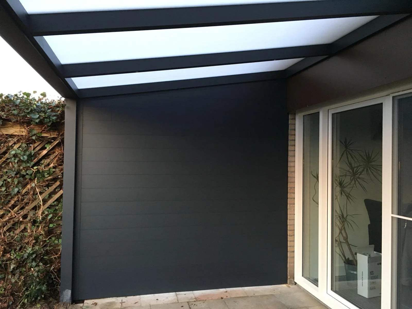 An exterior patio area with a black wall, glass sliding door, and a roof with translucent panels.