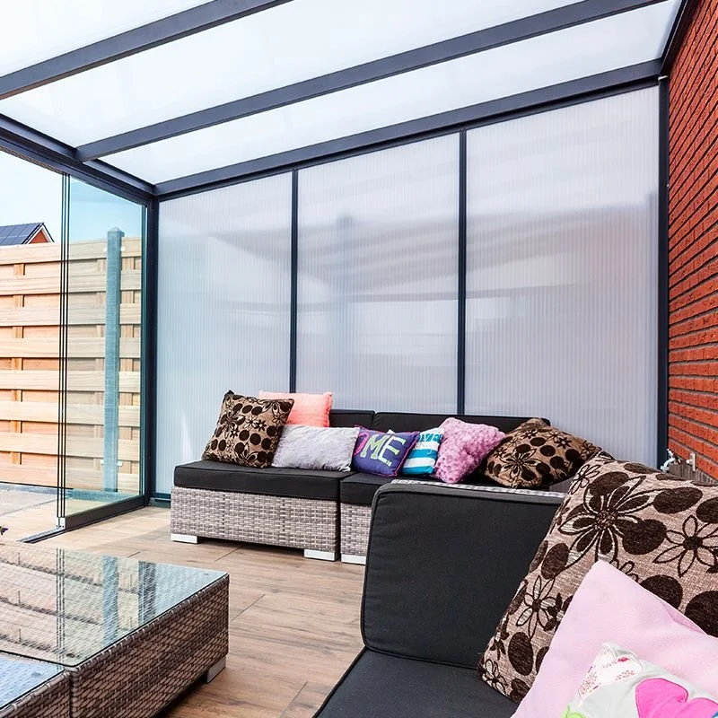 A sunroom with glass walls and a view of a backyard, featuring a woven outdoor sofa with colorful and patterned cushions, a glass-top coffee table, and a brick wall on the right.