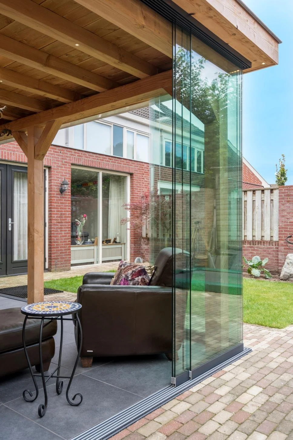An outdoor patio with a wooden pergola, black leather armchairs, a small decorative side table, and a glass wall separating the patio from a green yard with a brick house in the background.