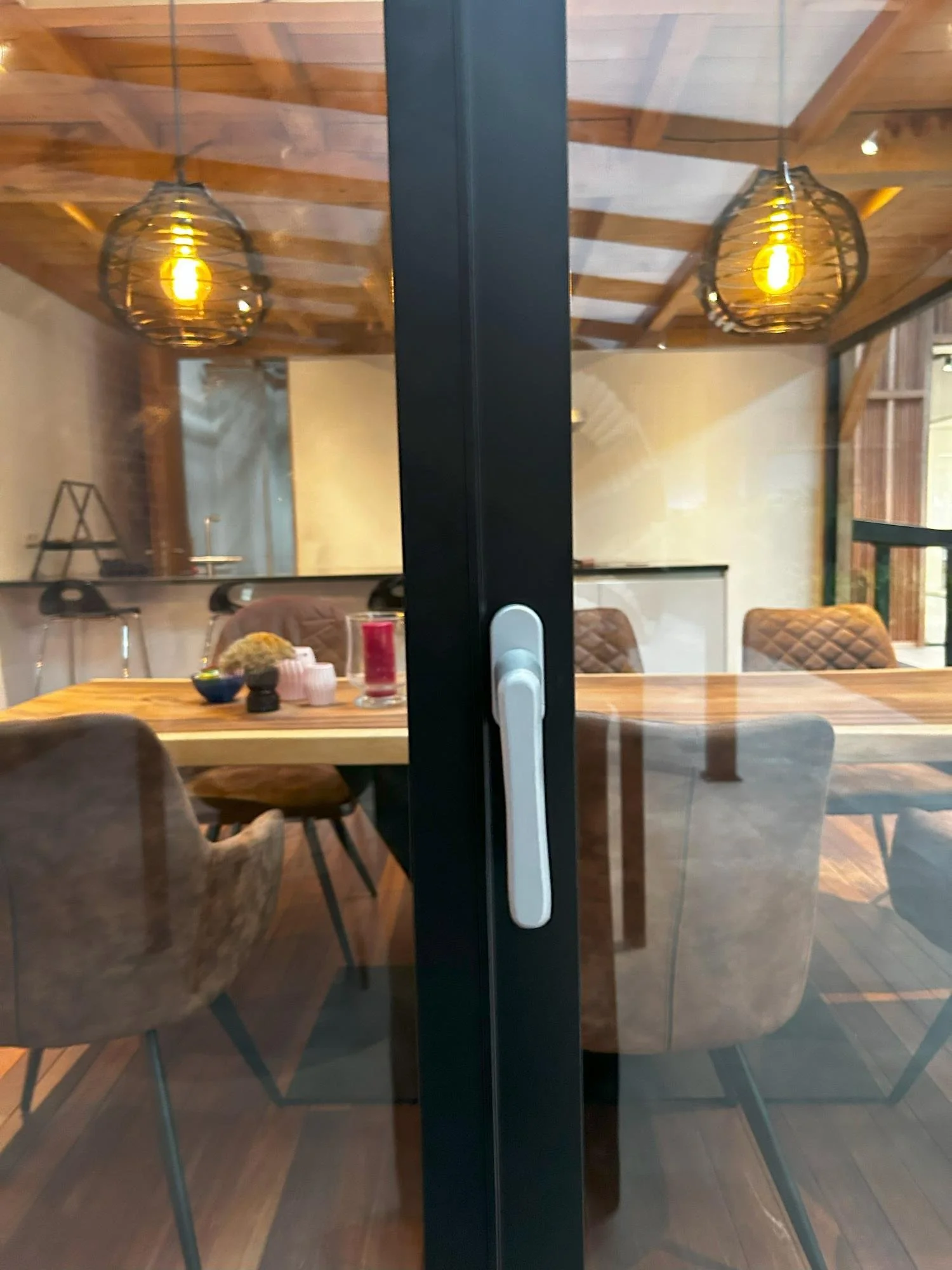 Interior view of a dining room seen through a glass door with a black frame and white handle. The room has a wooden ceiling with exposed beams, two hanging pendant lights, a wooden dining table, and several upholstered chairs. The table has some deco