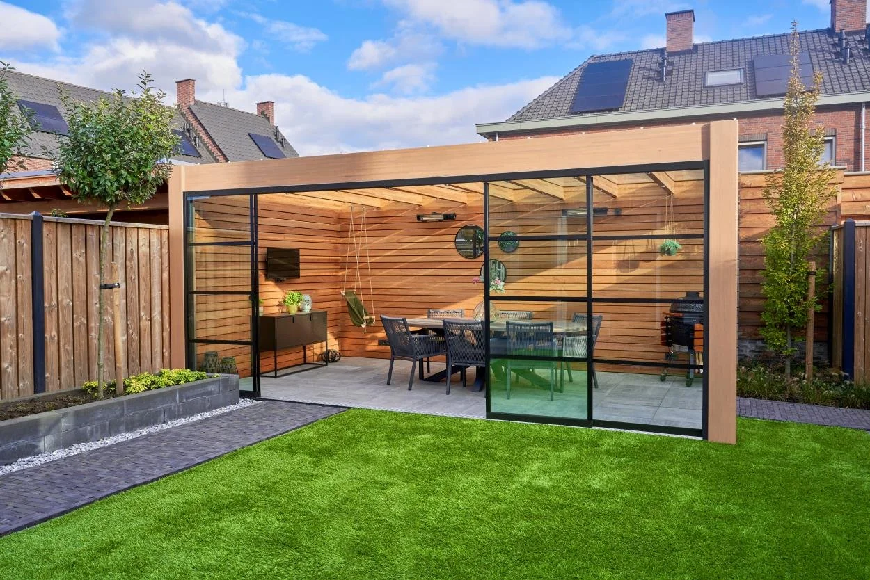 A backyard with a modern wooden and glass outdoor pavilion, outdoor dining table with six chairs, a barbecue grill, and a green lawn under a partly cloudy sky.