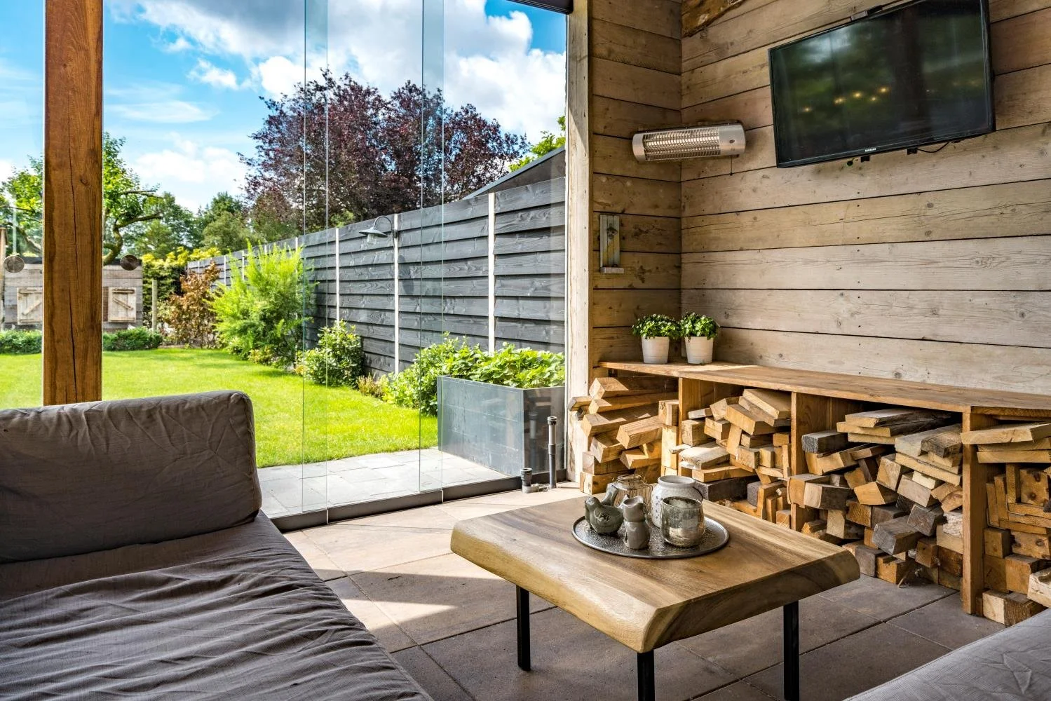 Interior of a cozy room with wooden walls and a sliding glass door leading to a lush green yard with trees and shrubs. The room features a wooden coffee table with decorative items, a gray fabric sofa, a mounted flat-screen TV, and built-in shelves f