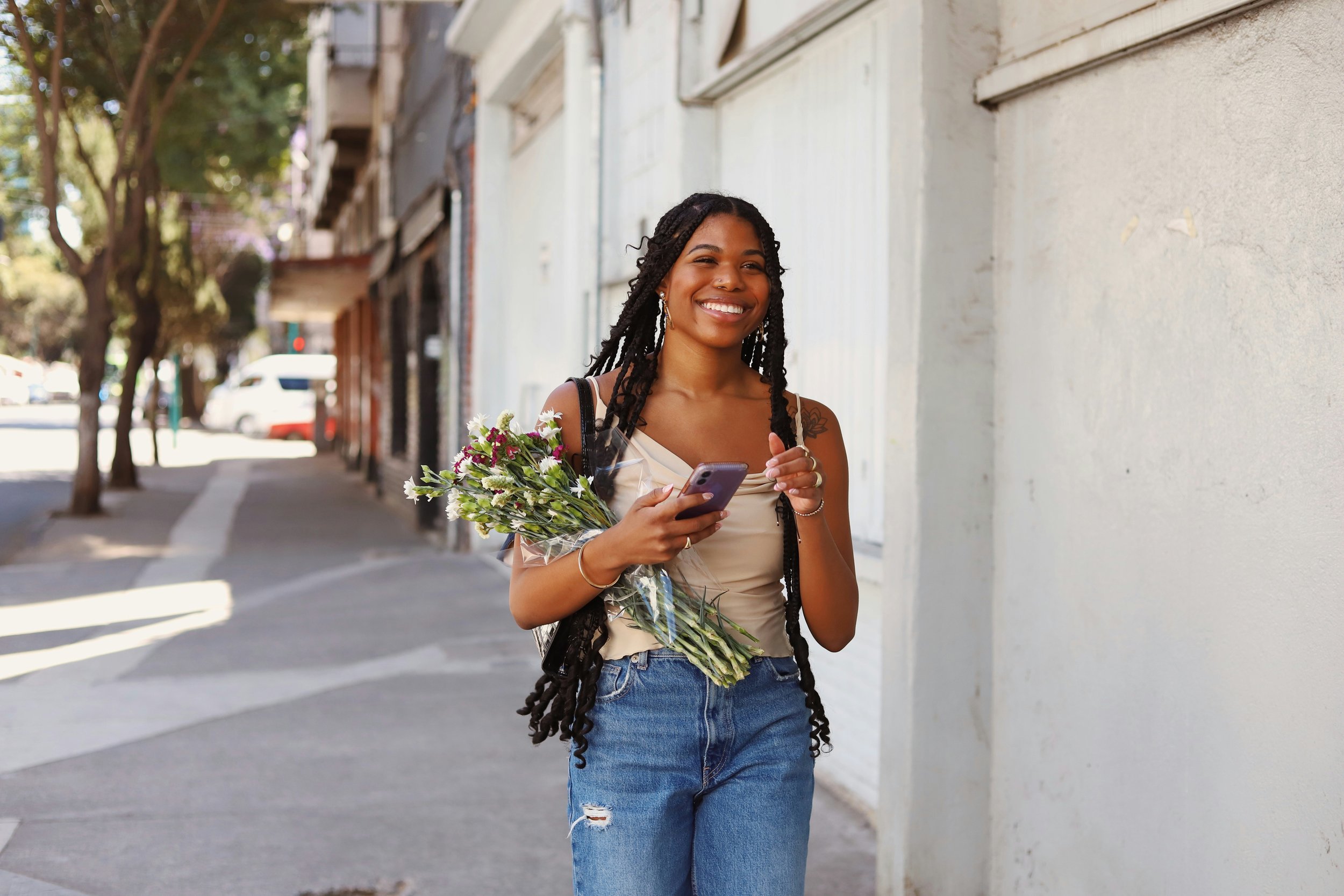A young woman with long dreadlocks smiling while holding a bouquet of flowers and a smartphone on a city sidewalk.