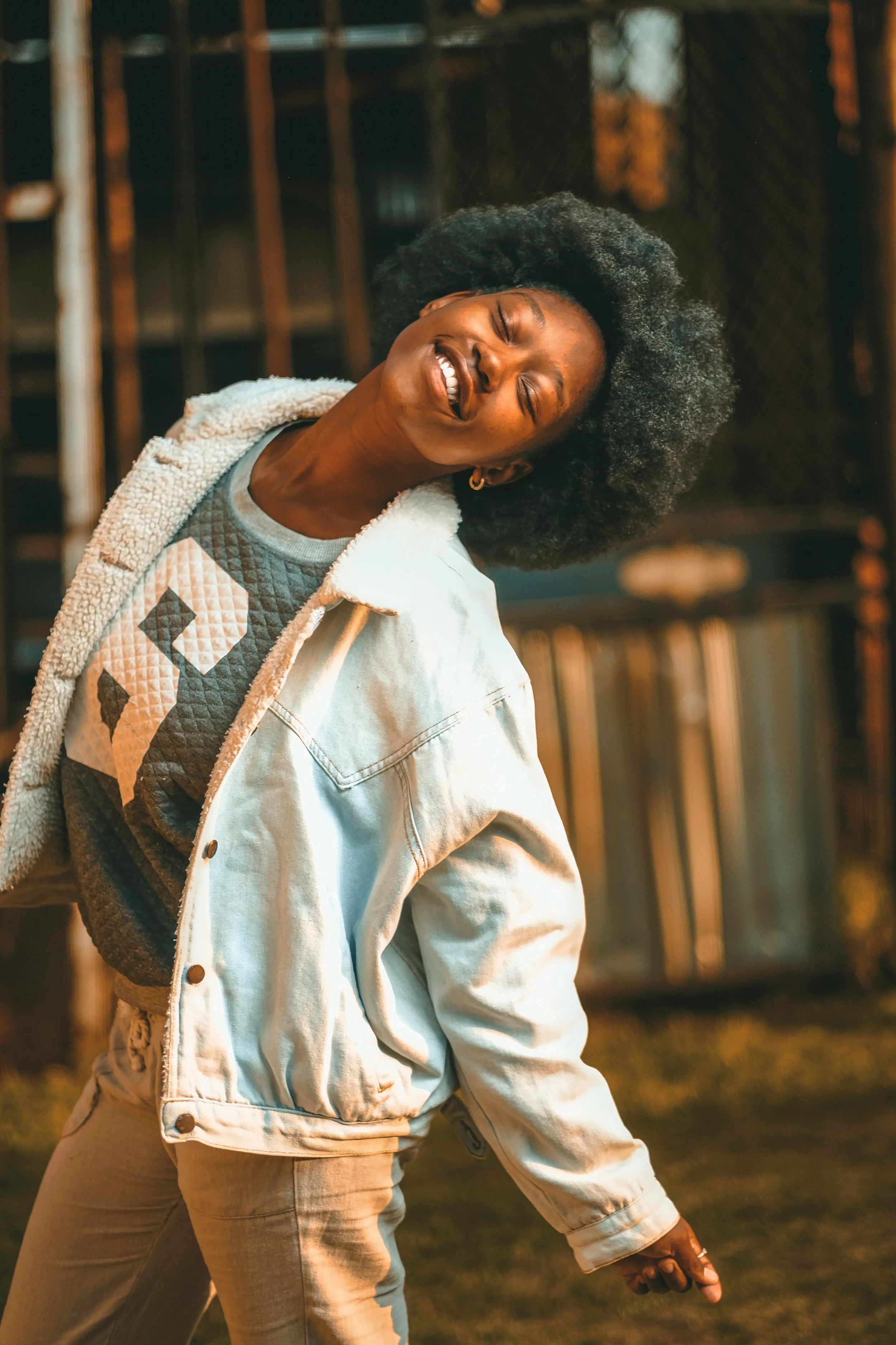 A woman with curly black hair, smiling and laughing, wearing a denim jacket over a patterned sweater, standing outdoors at sunset.