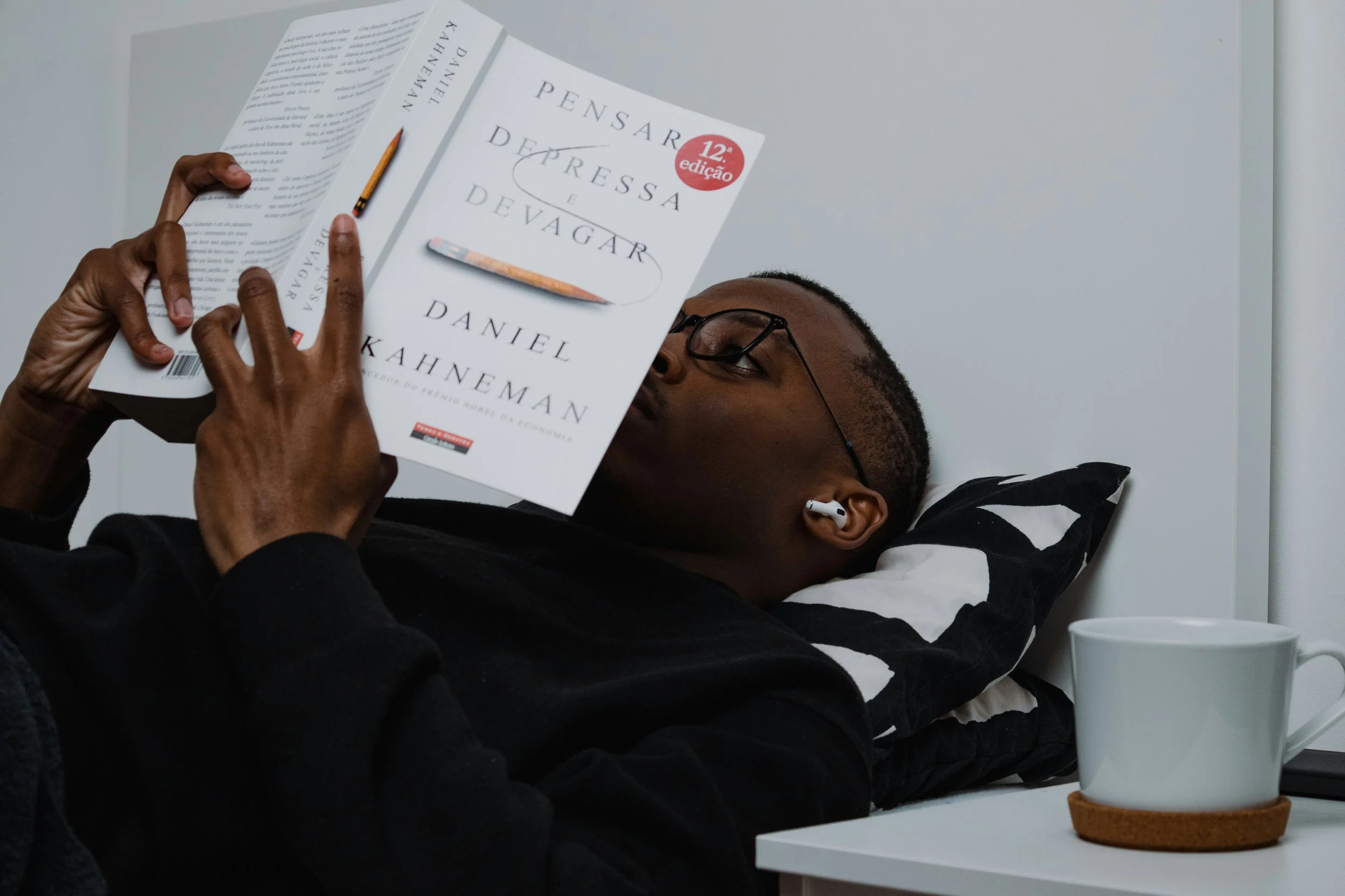 A person lying in bed reading a book titled 'Pensar a depressa e devagar,' with a coffee mug on the bedside table.