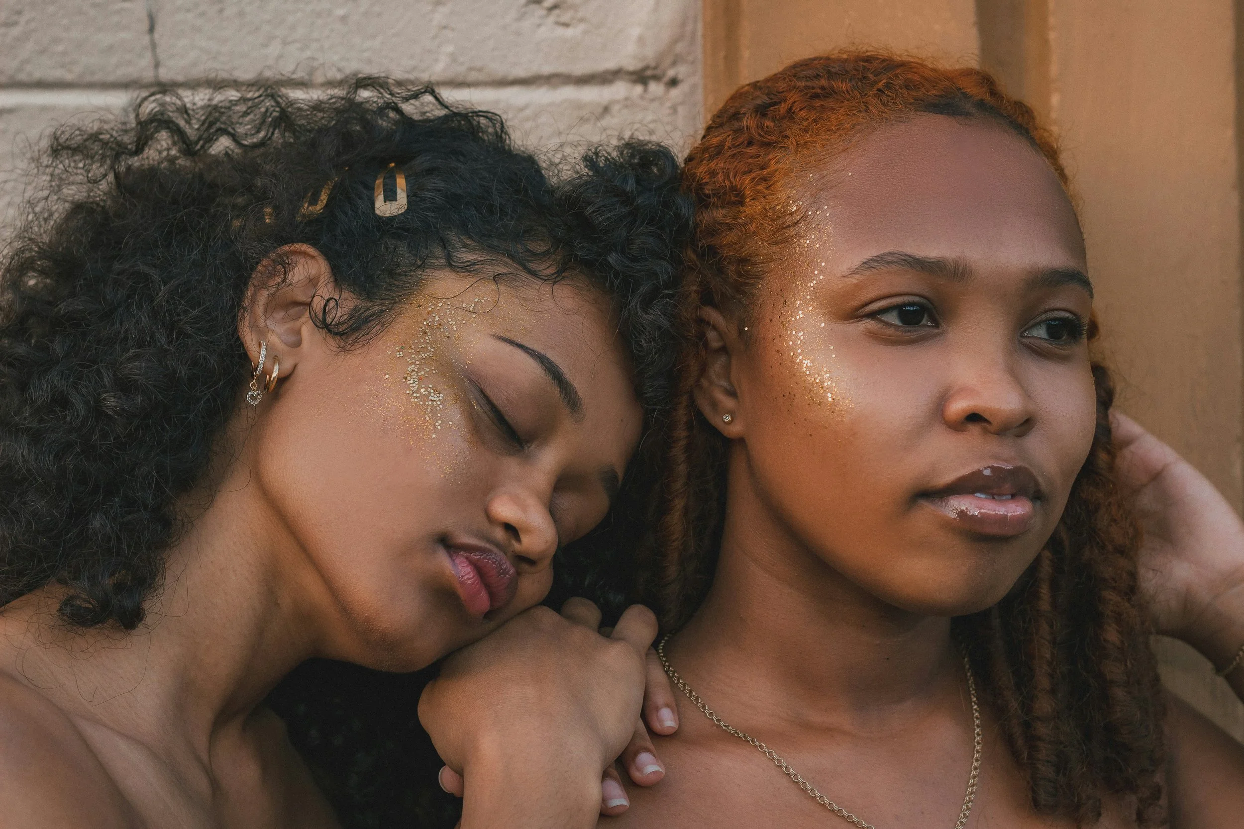 Two young women with gold glitter on their faces and wearing jewelry, resting against a brick wall, one with closed eyes and the other looking forward.
