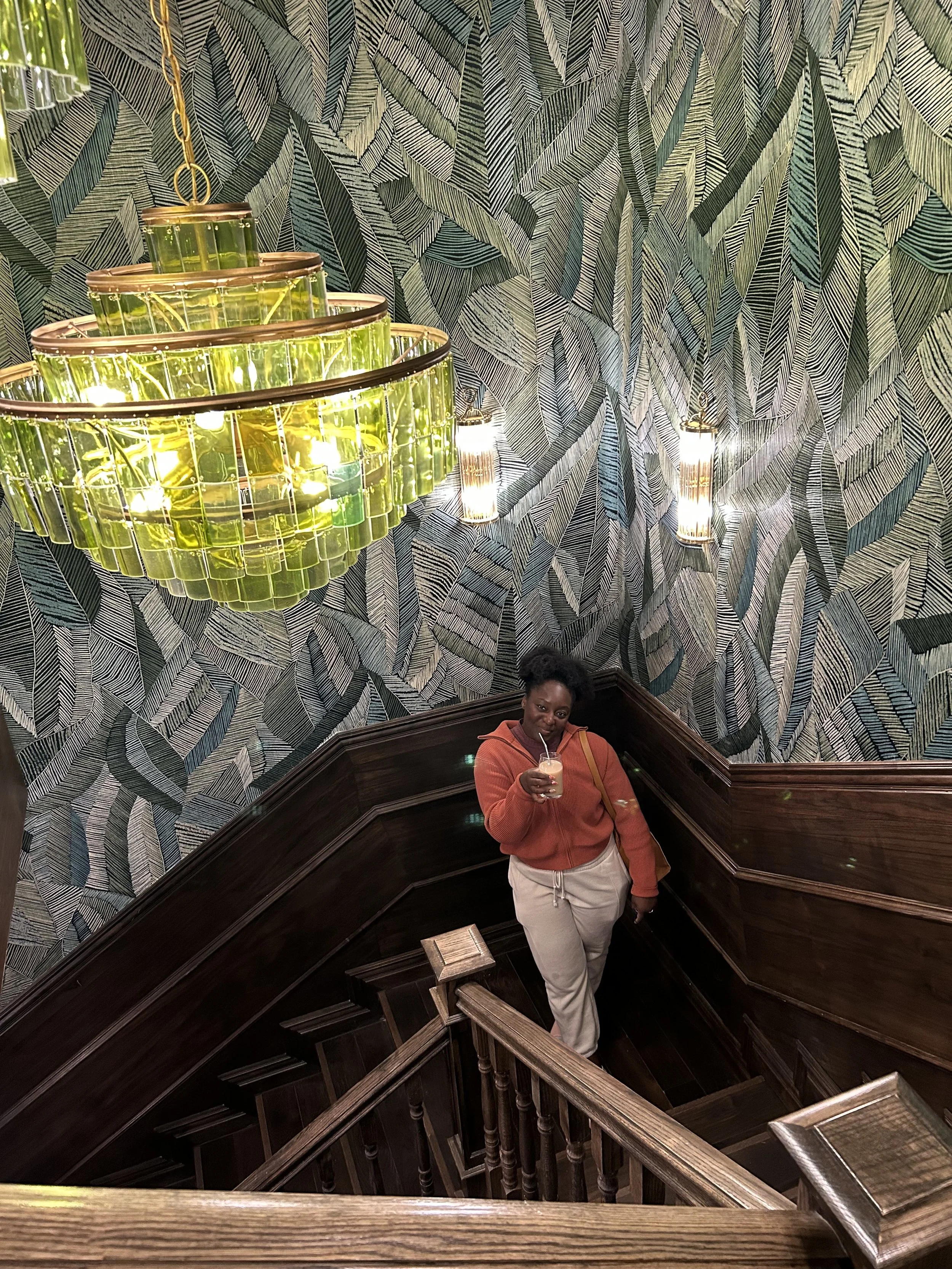 A woman standing on a staircase in a themed room with leafy wallpaper, holding a drink and looking up at the camera. A large green chandelier and wall sconces are visible overhead.