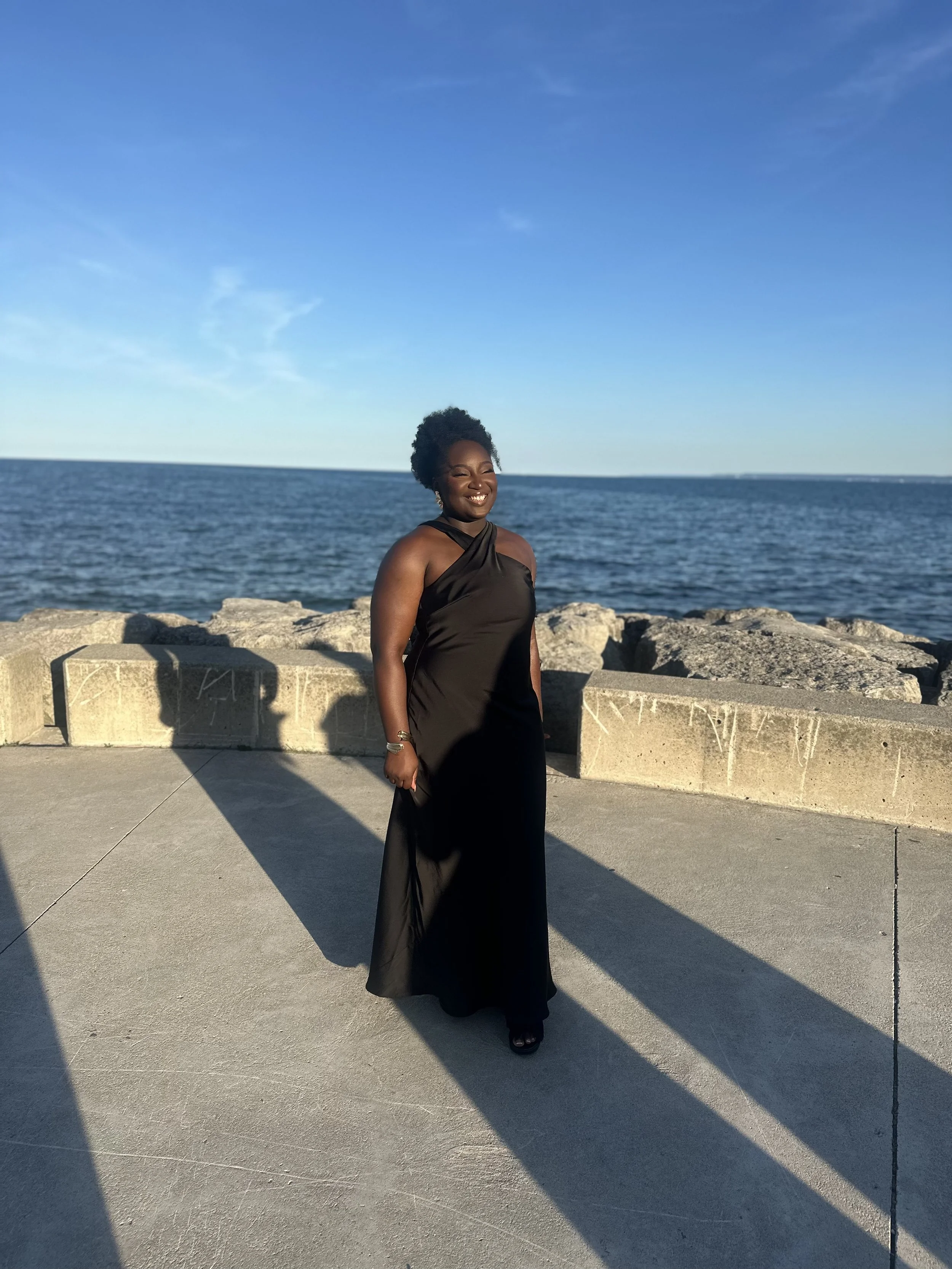 A woman in a black dress standing on a concrete platform near the ocean with a stone barrier, smiling as the sun sets, casting long shadows.