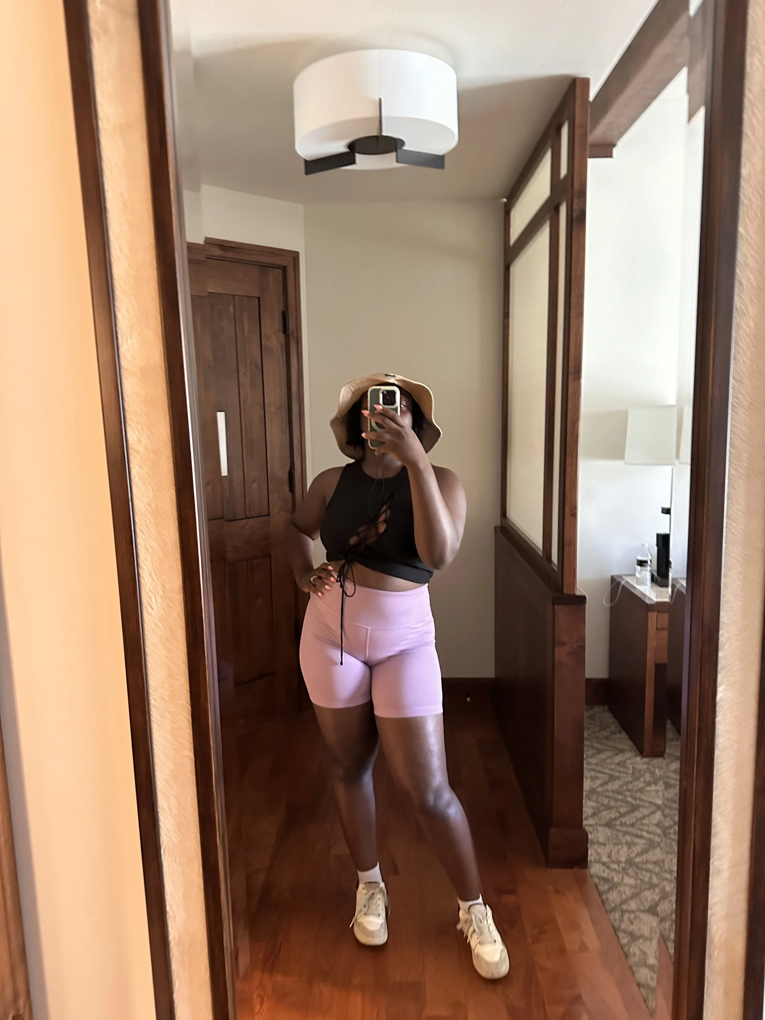 A woman wearing a wide-brimmed tan hat, black sleeveless crop top, pink shorts, white sneakers, takes a mirror selfie in a hotel room with wooden furniture and a ceiling-mounted light fixture.