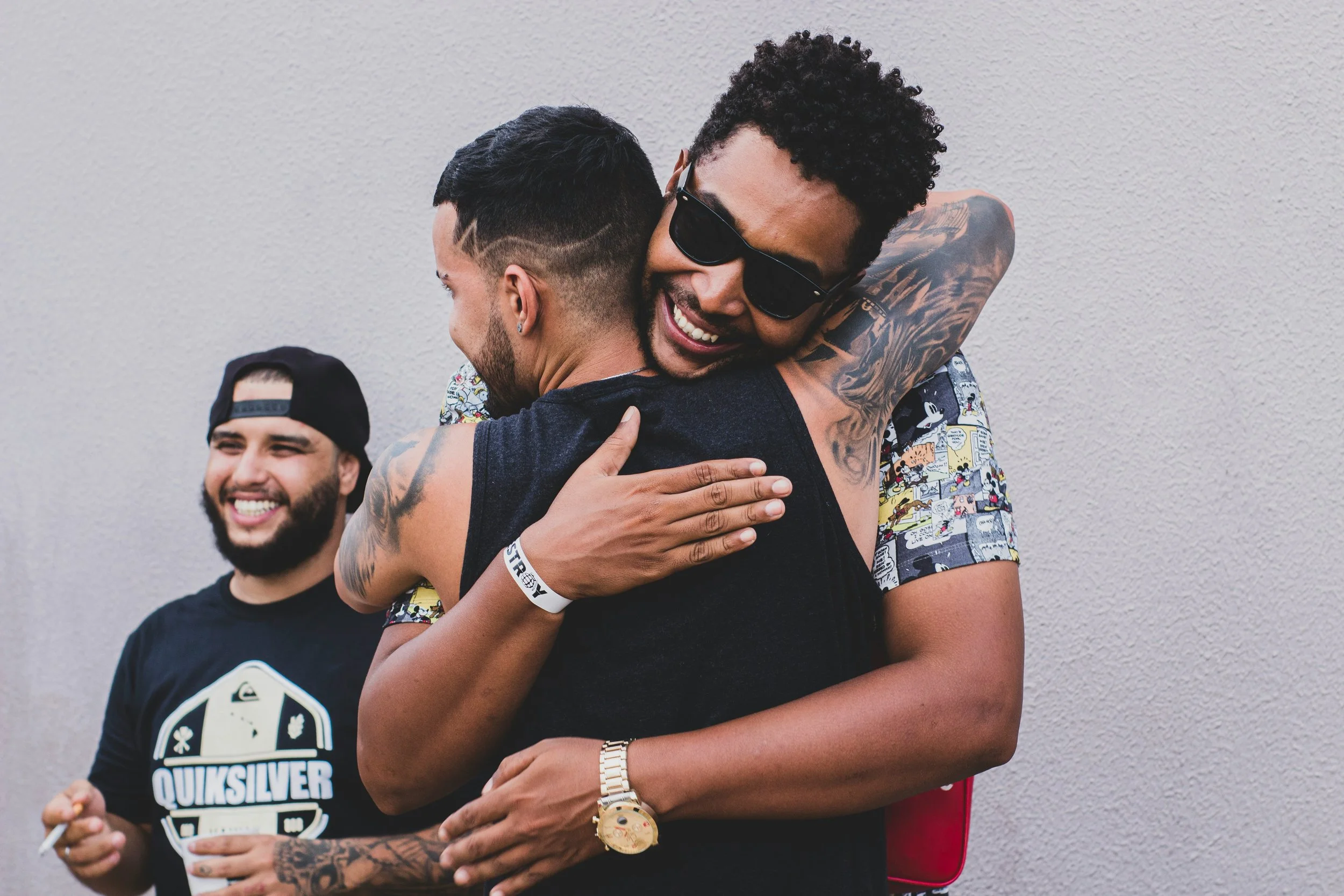 Three young men are sharing a joyful, friendly moment. One man is hugging another with a broad smile, wearing sunglasses and a short-sleeved T-shirt with a colorful cartoon print. The man being hugged has tattoos on his arms and is wearing a sleeveless shirt. The third man, in the background, is smiling and wearing a black quiksilver T-shirt, a black cap backwards, and holding a cigarette.