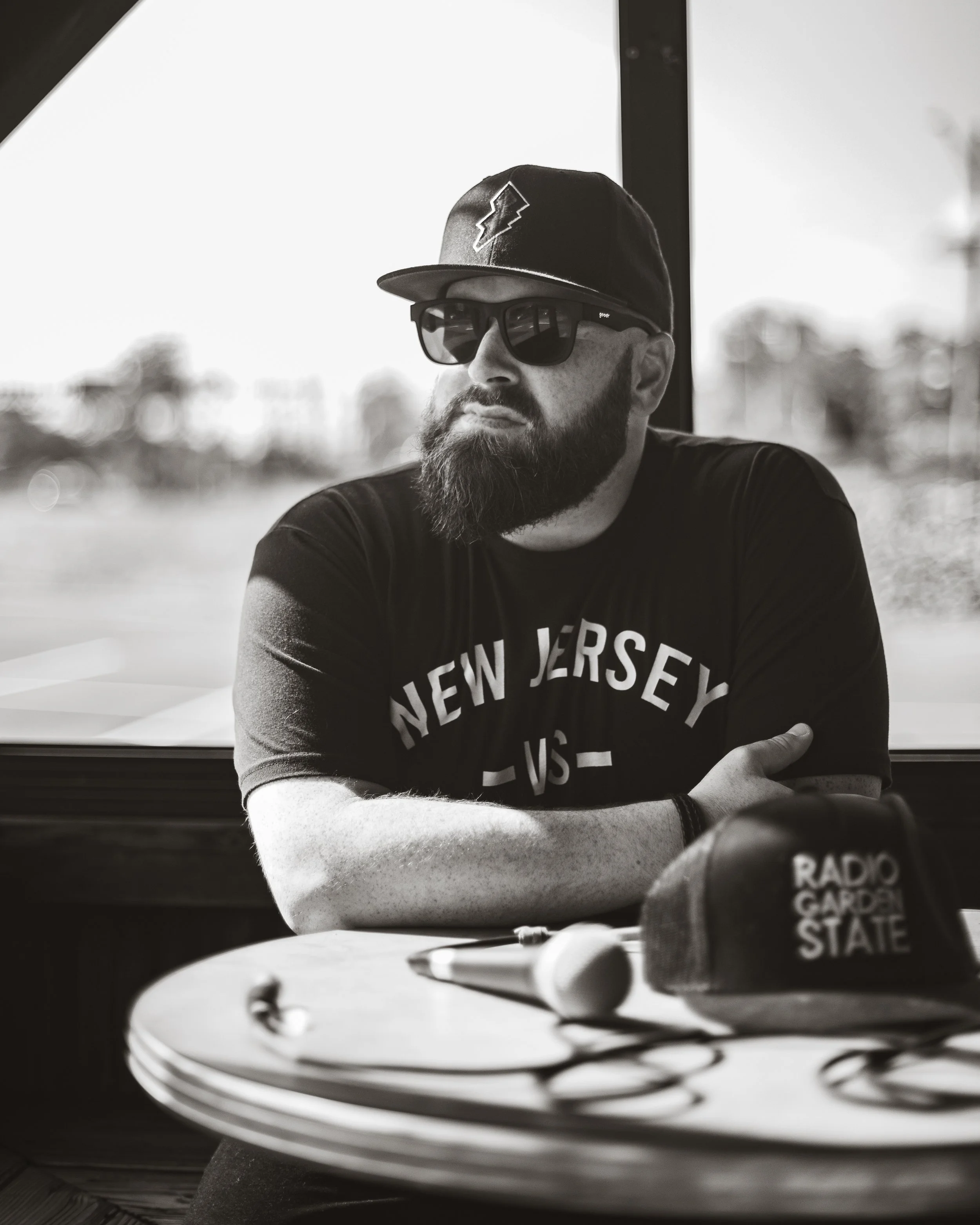 A man with a beard wearing sunglasses, a baseball cap, and a t-shirt with 'New Jersey' printed on it, sitting at a table with radio station equipment in front of him, in a brightly lit setting with blurred outdoor background.