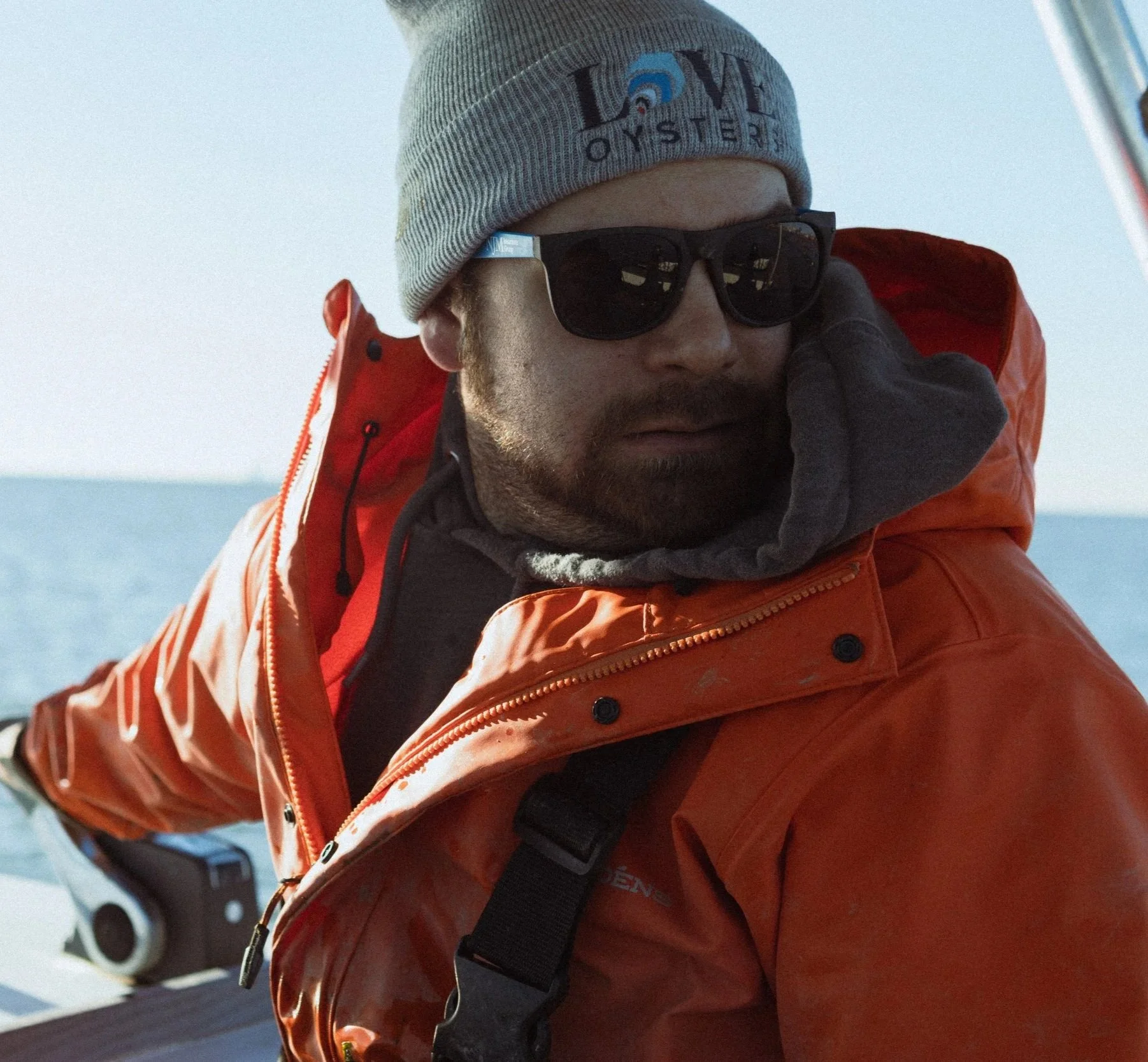 Man wearing a gray beanie, black sunglasses, and an orange waterproof jacket on a boat with the ocean in the background.