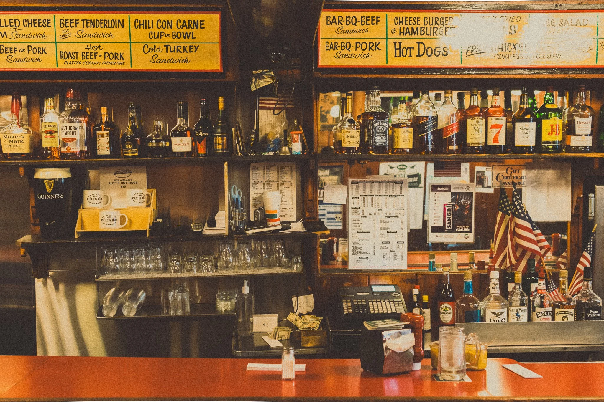 Bar counter with various liquor bottles, American flags, glassware, and menus in a dimly lit bar.