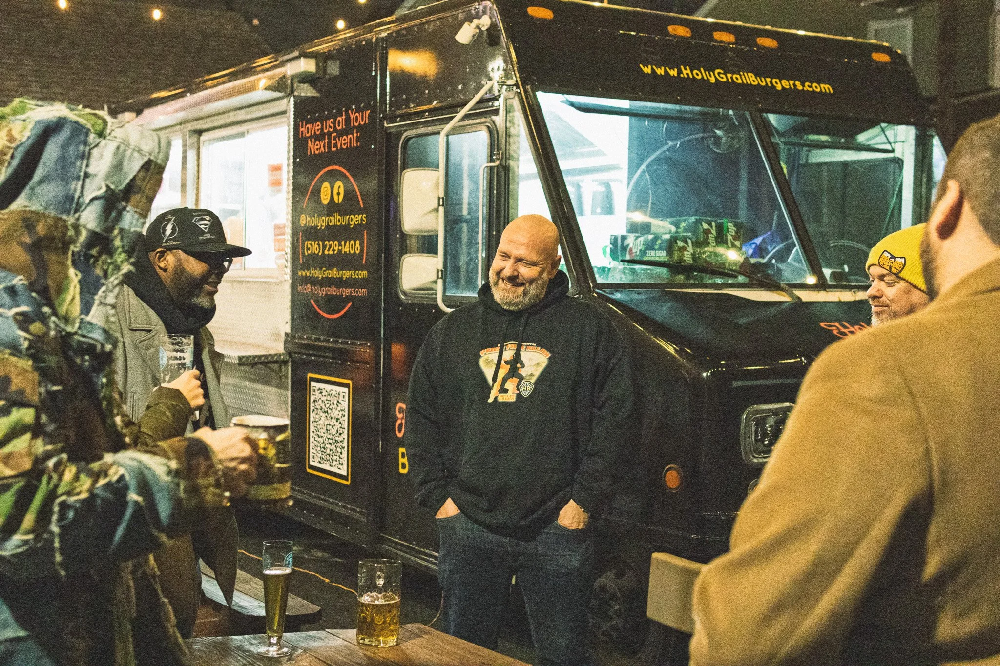 Group of men socializing outside at night around a hot dog food truck with drinks on a wooden table.