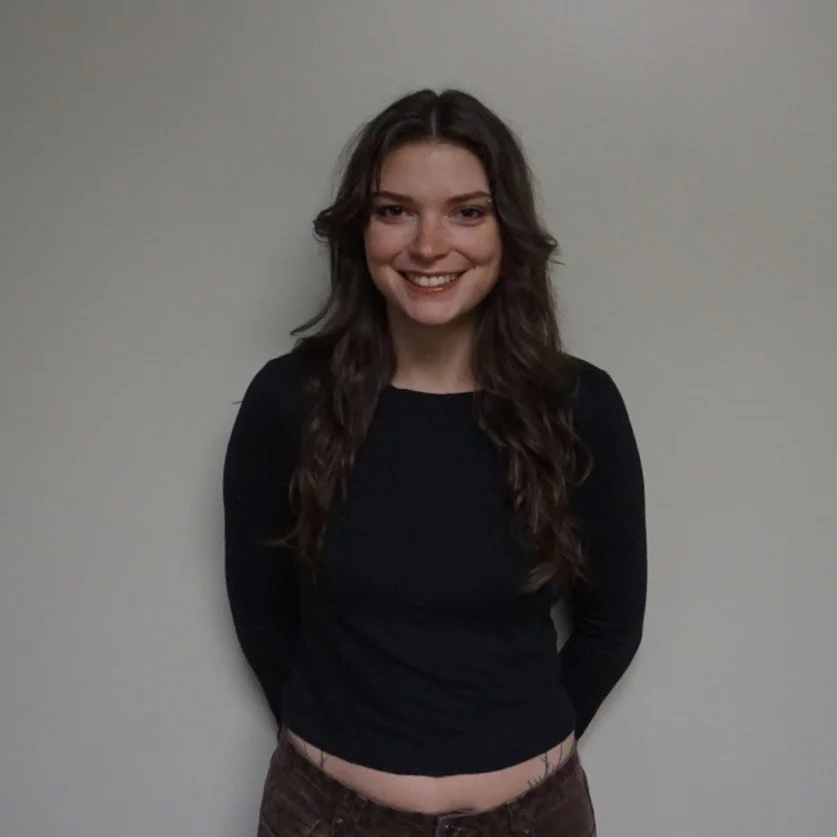 A young woman with long brown hair, smiling, wearing a black long-sleeve shirt and brown pants standing against a plain white wall.
