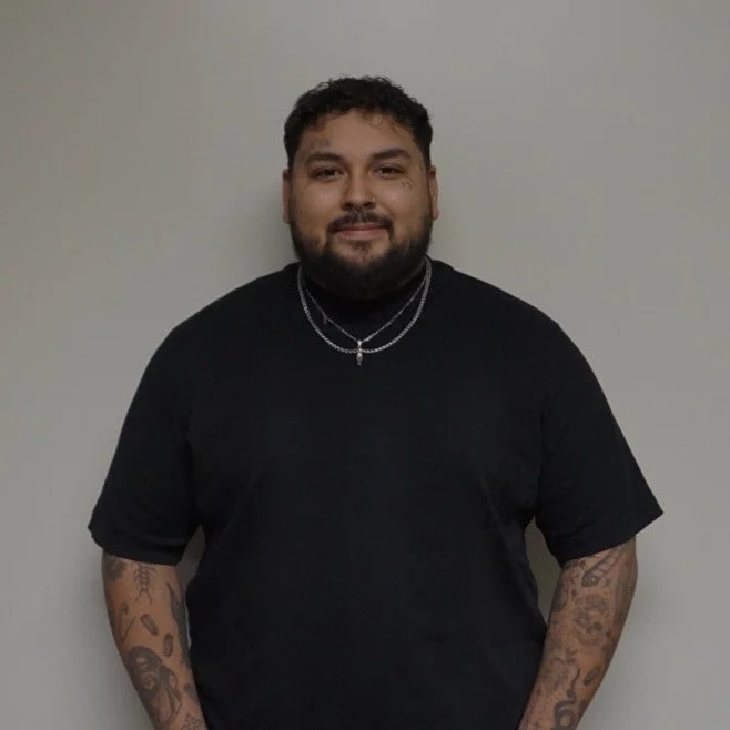 A man with dark curly hair, a beard, and tattoos on his arms, wearing a black T-shirt and silver jewelry, standing against a plain light gray wall.