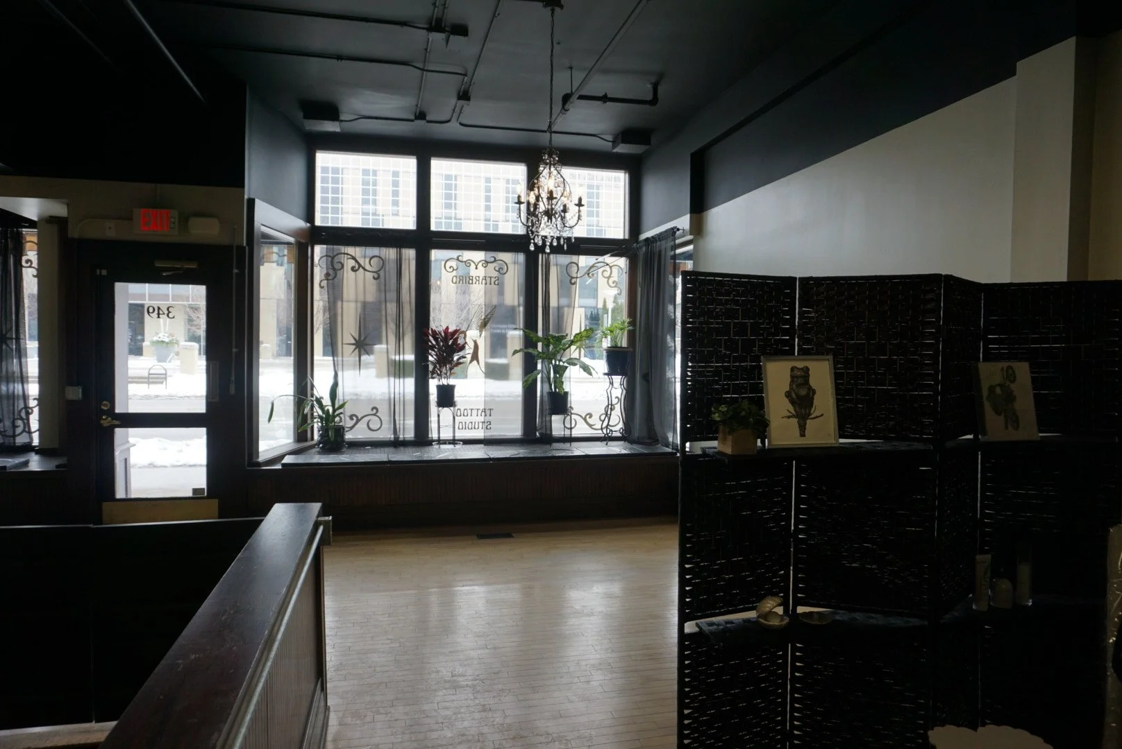 Interior of a dimly lit boutique or studio with large front windows, plants, a chandelier, and decorative items on dark partitions.