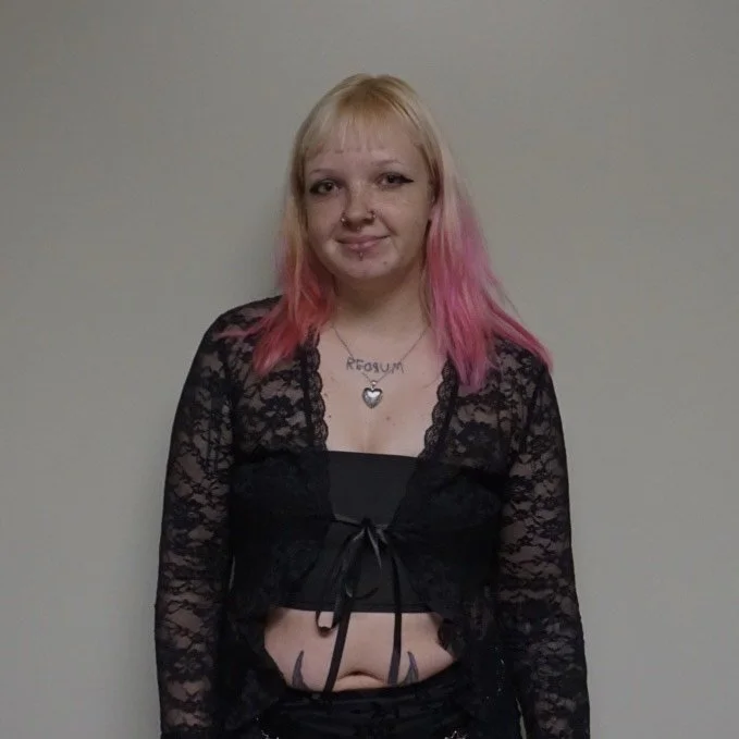 A young woman with blond hair with pink tips, wearing a black lace top and a necklace with a heart-shaped pendant, standing against a plain wall.