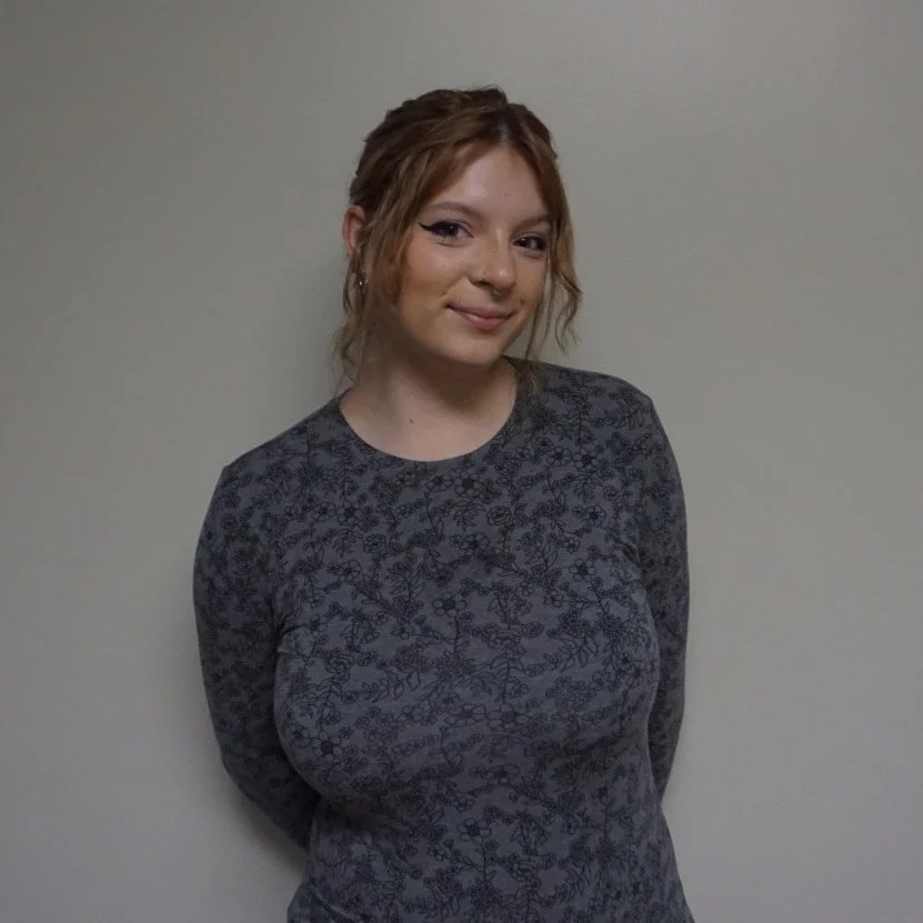 A young woman with reddish-brown hair styled in loose waves, wearing a patterned long-sleeve shirt, standing against a plain off-white wall, smiling gently at the camera.