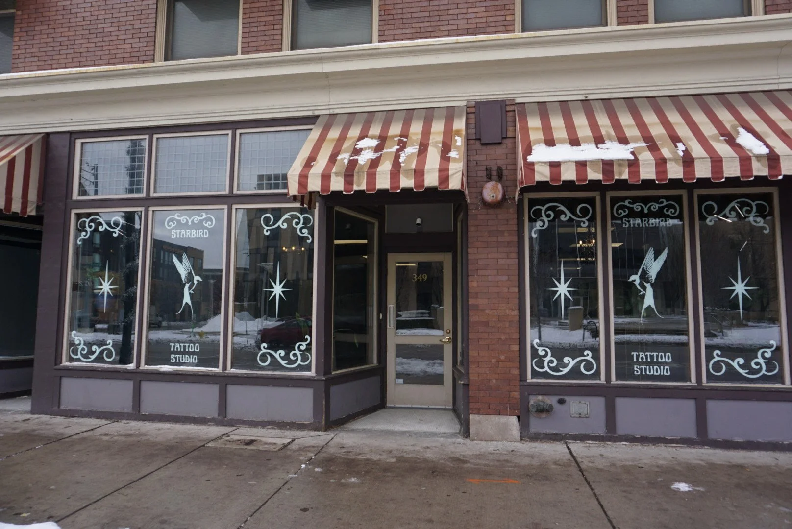 Exterior view of a tattoo studio named Starbird with large window decals showing a fairy holding a star, snow on awnings and ground, brick building facade, and the address 349 on the door.